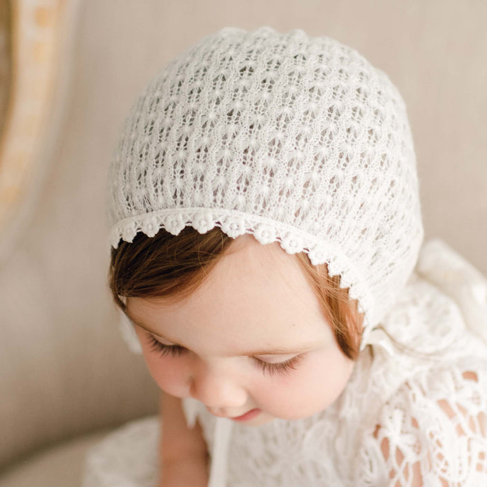 Close-up view of a baby girl in the Lola Christening Knit Bonnet, crafted from soft light ivory acrylic knit, featuring delicate cotton lace trim and silk ribbon ties. She is gazing down away from the camera.
