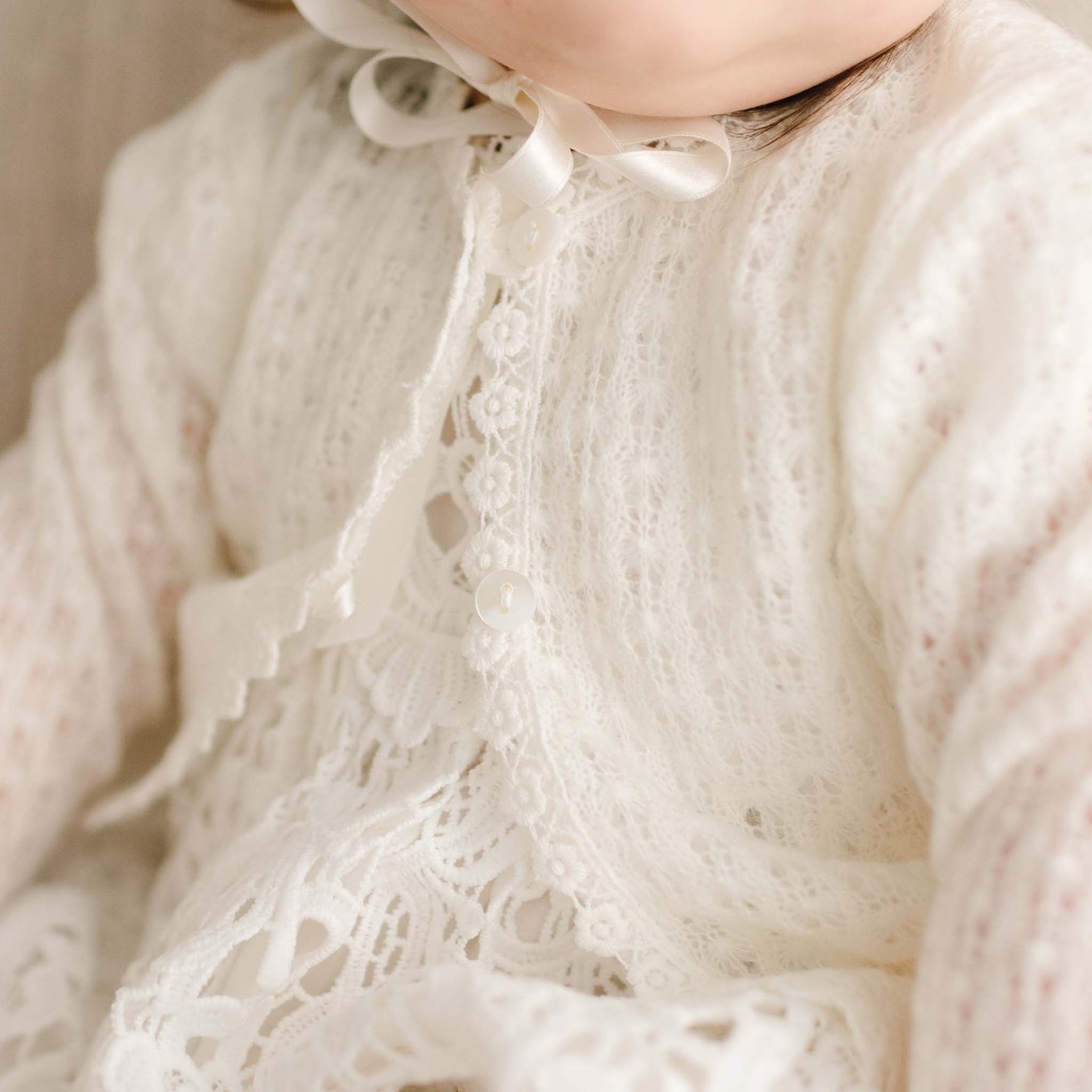 Close-up of the Lola Knit Christening Sweater, featuring soft light ivory knit fabric, button closures, and delicate lace trim, layered over a lace christening gown.