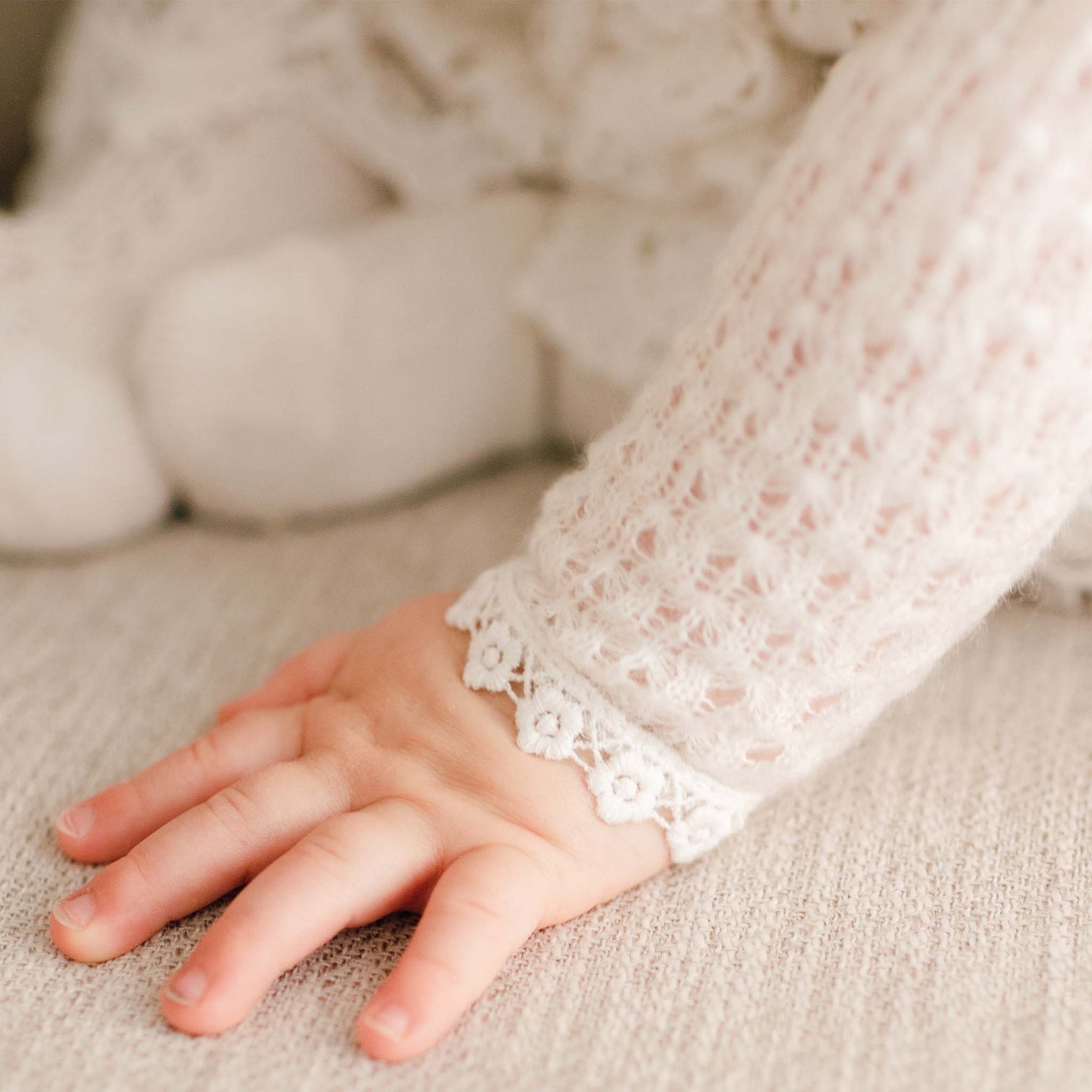 Close-up of a baby’s hand resting on fabric, wearing the Lola Christening Knit Sweater with intricate light ivory knit fabric and lace-trimmed cuffs.