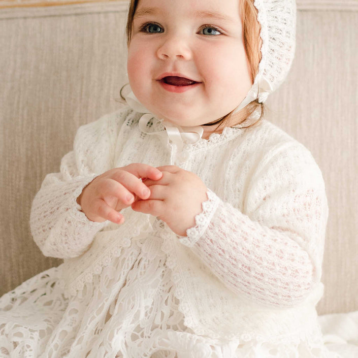 Baby girl wearing the Lola Knit Sweater over the Lola Christening Gown. The sweater is made from soft, breathable light ivory knit acrylic, with button closures and delicate ivory trim.