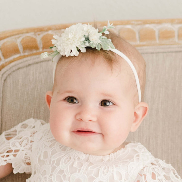 Baby girl wearing the Lola Christening Flower Headband, featuring realistic cream flowers and soft green leaves, paired with a lace christening outfit.