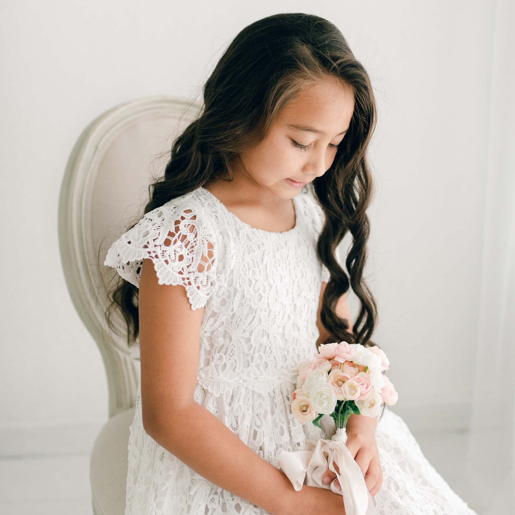 A girl in the Lola Girls Lace Dress holds a small bouquet, looking downward with long curled hair cascading over her shoulder.