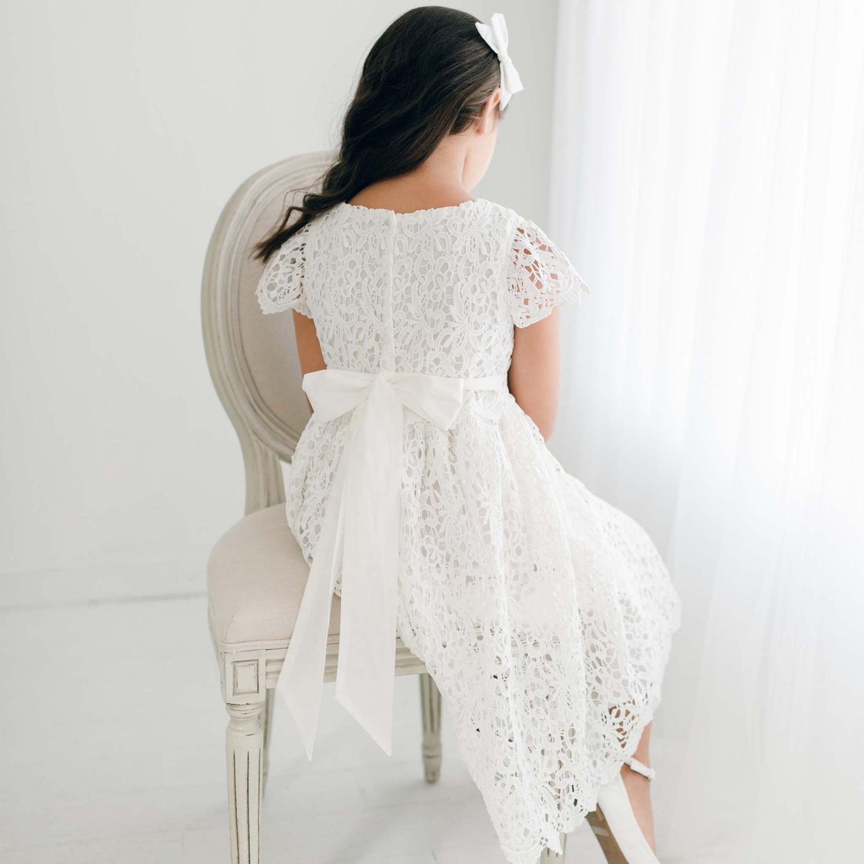 The back of the Lola Girls Lace Dress is displayed as a girl sits on a chair, highlighting the delicate lace design and a satin sash tied in a bow.
