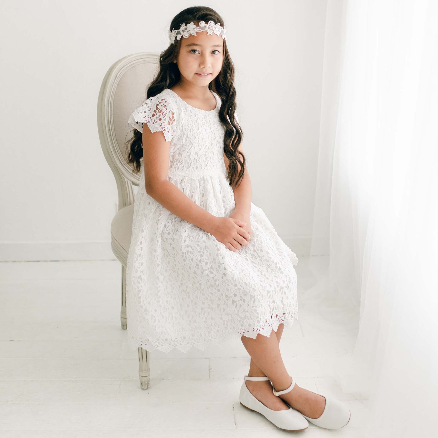 A girl sits gracefully on a vintage-style chair, wearing the Lola Girls Lace Dress with a lace headband, hands gently folded in her lap.