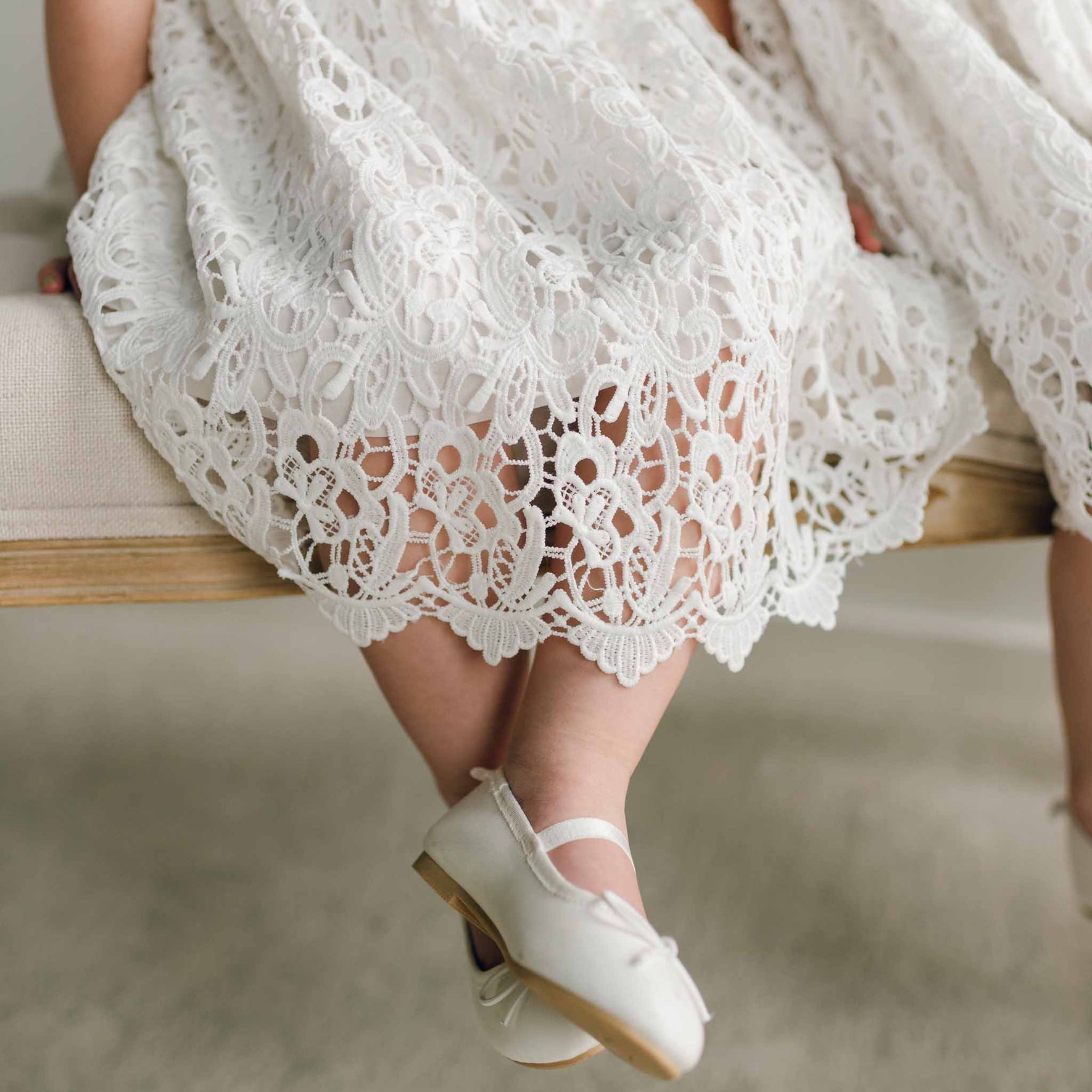Close-up of the Lola Girls Lace Dress hem, showcasing the intricate lace pattern as a child's feet dangle over the edge of a bench in white Mary Jane shoes.
