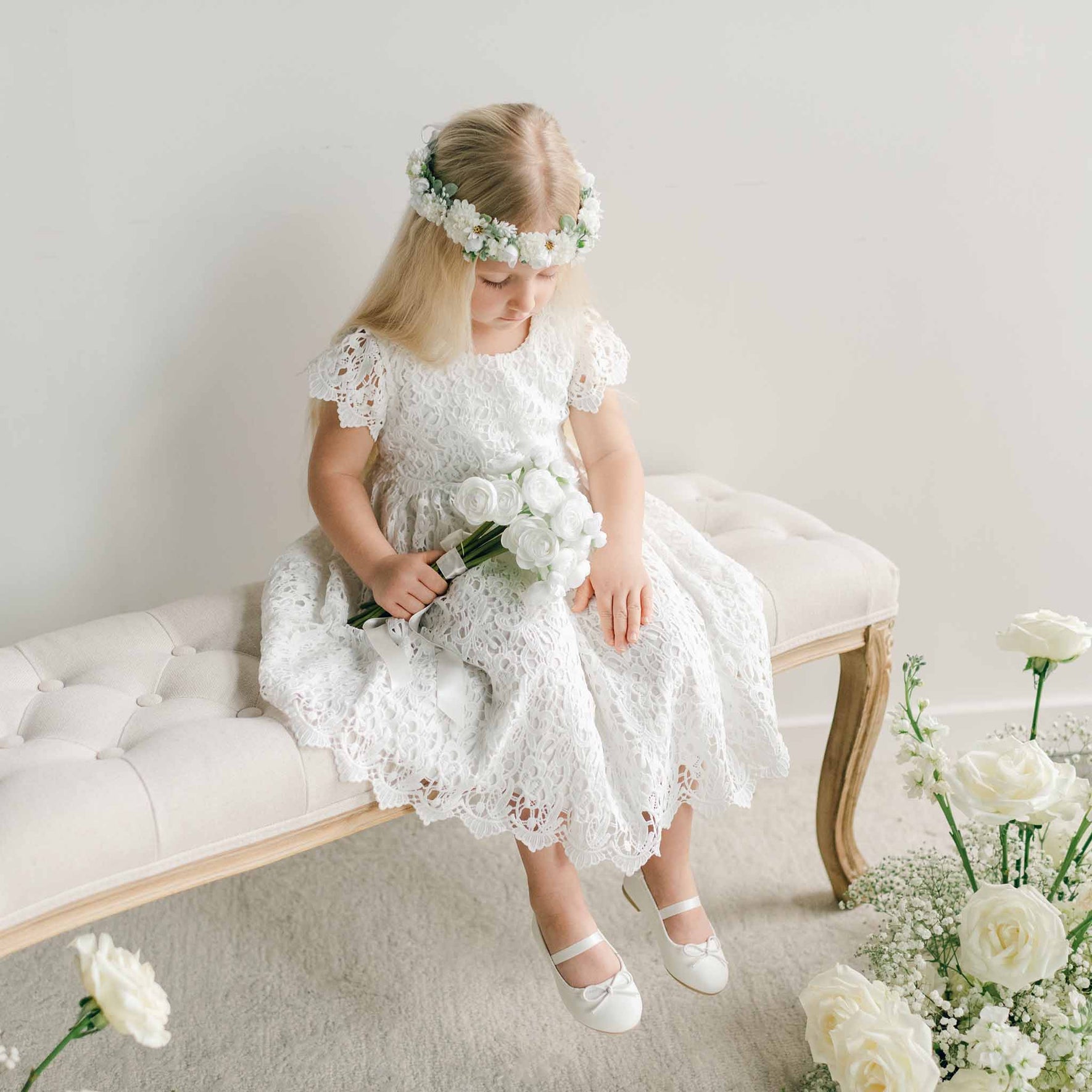 A young girl in the Lola Girls Lace Dress sits on a tufted bench, looking down at her bouquet of white flowers, with soft floral arrangements around her.