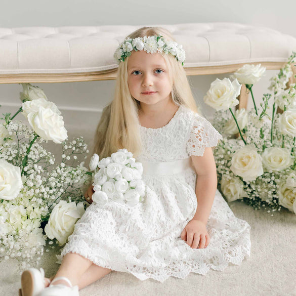 A young girl sits on the floor surrounded by white roses, wearing the Lola Girls Lace Dress with a floral headband and holding a bouquet of white flowers.