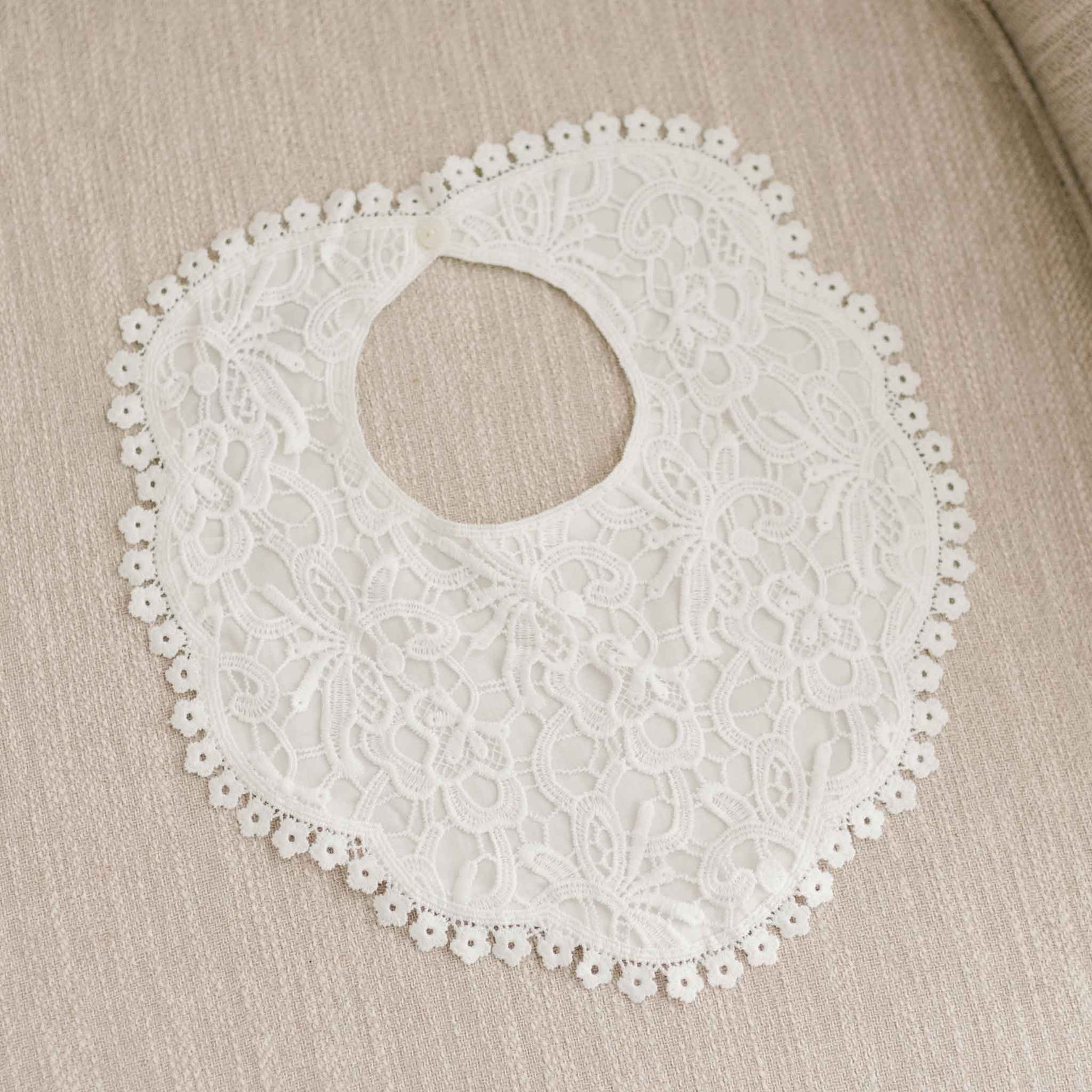 Top-down view of the Lola Lace Christening Bib on linen fabric, featuring a delicate lace overlay, cotton lace trim, and a button closure at the back,
