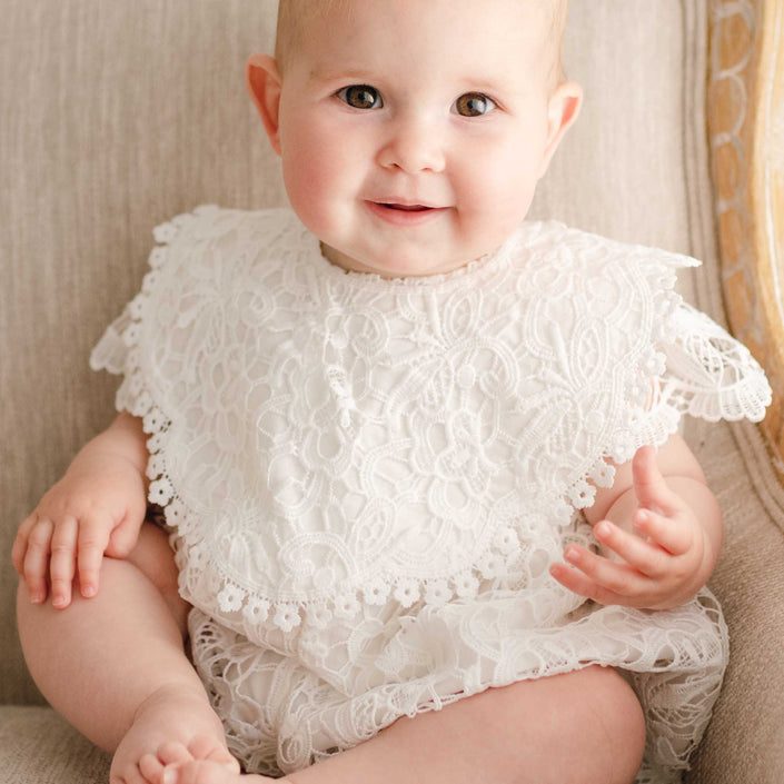 Newborn baby girl wearing the Lola Lace Christening Bib, featuring a delicate lace overlay and cotton lace accents, over a lace romper, looking at the camera with a slight smile.