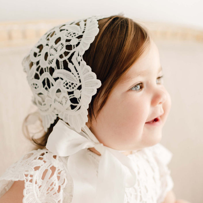 Baby girl with brown hair smiles softly, wearing the Lola Lace Christening Bonnet and matching lace gown, with intricate lace details highlighted by natural light.