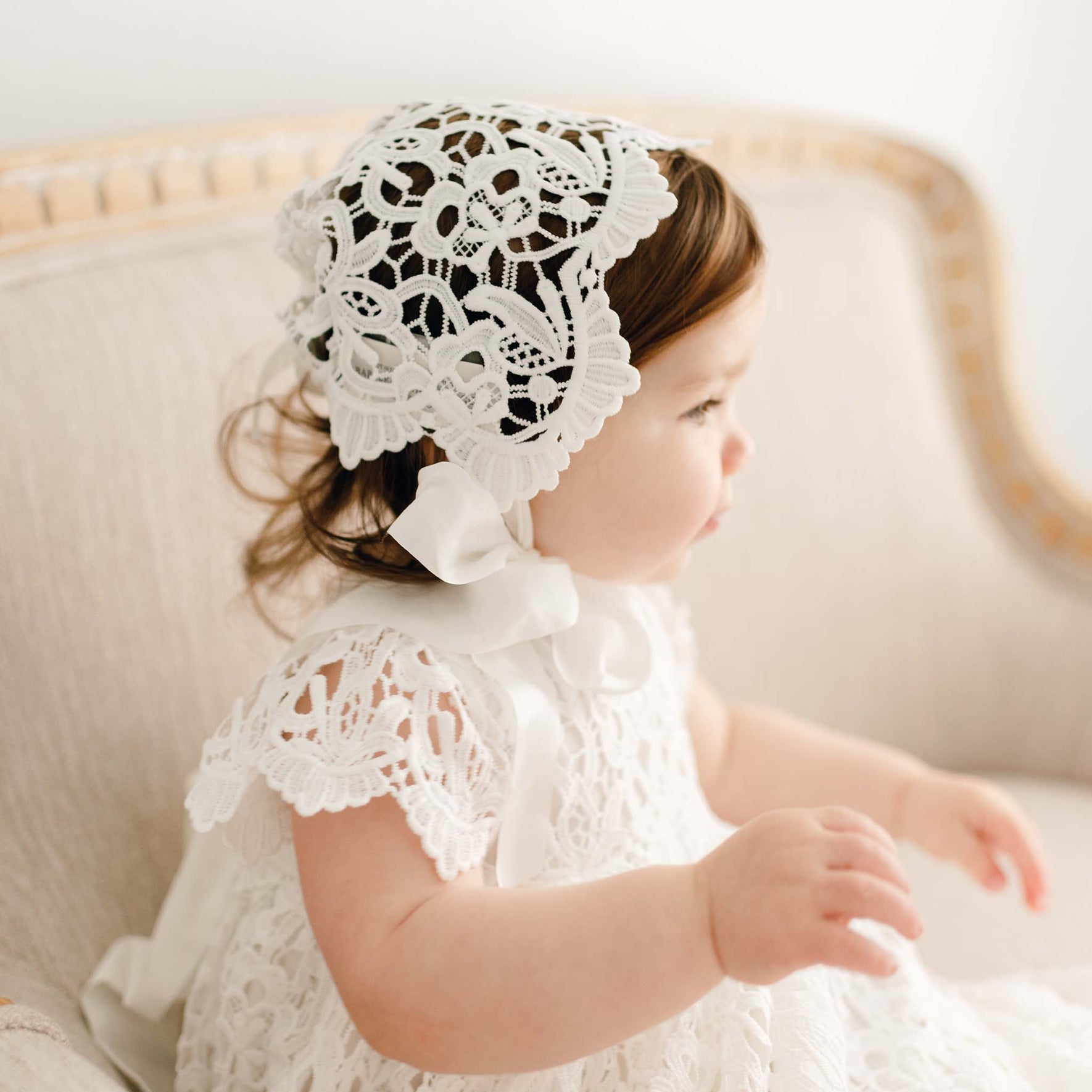 Baby girl wearing the Lola Lace Bonnet, a delicate white lace christening bonnet with satin ribbon ties.
