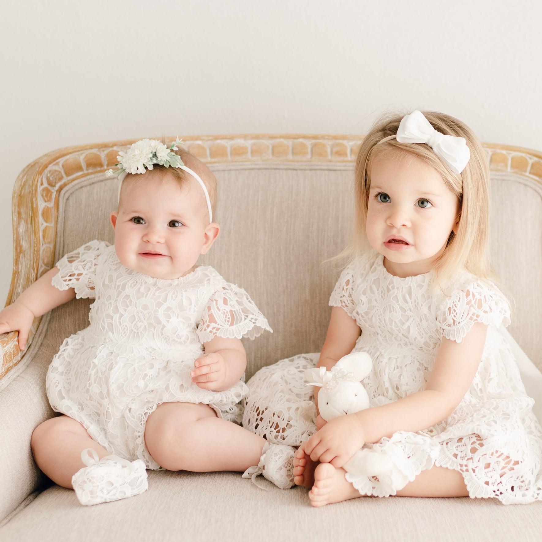 Two baby girls in the Lola Christening collection: the baby on the left is wearing the Lola Christening Romper with a coordinating floral headband, and the baby on the right is dressed in the Lola Christening Dress with a matching white bow headband.