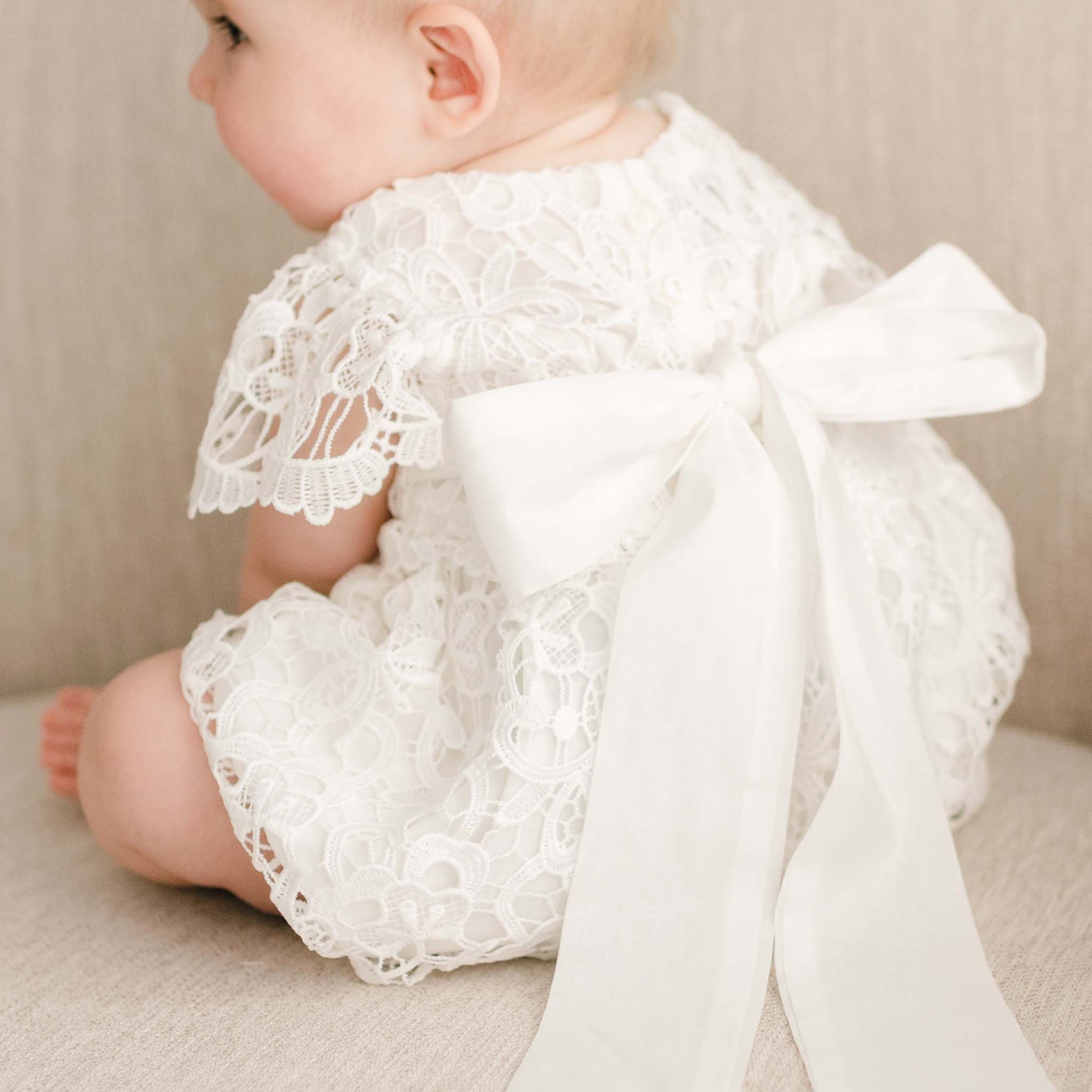 Back view of a baby girl sitting on a linen chair, wearing the Lola Lace Christening Romper, featuring a large sash tie fastened into a bow.