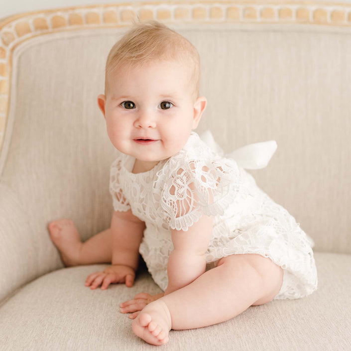 Baby girl wearing the Lola Lace Christening Romper, showcasing delicate light ivory floral lace, seated on a linen-upholstered chair.