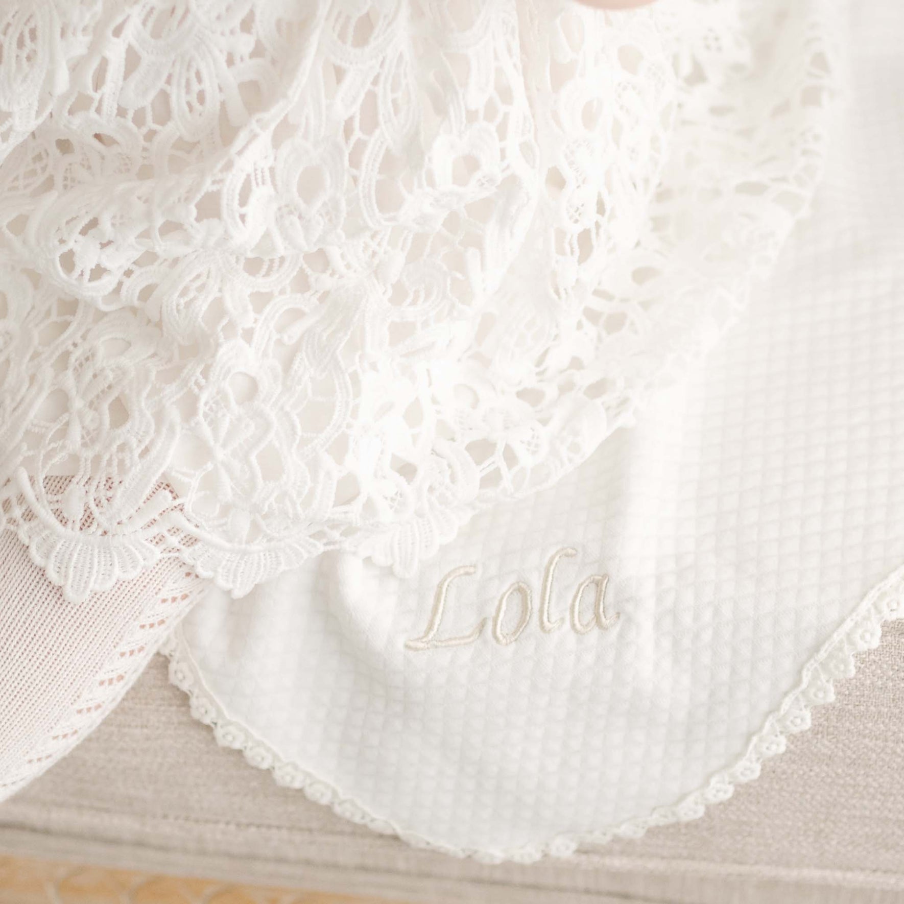 Close-up of the Lola Christening Personalized Blanket in light ivory cotton with lace trim, featuring 'Lola' embroidered on the corner, paired with a delicate lace christening gown.