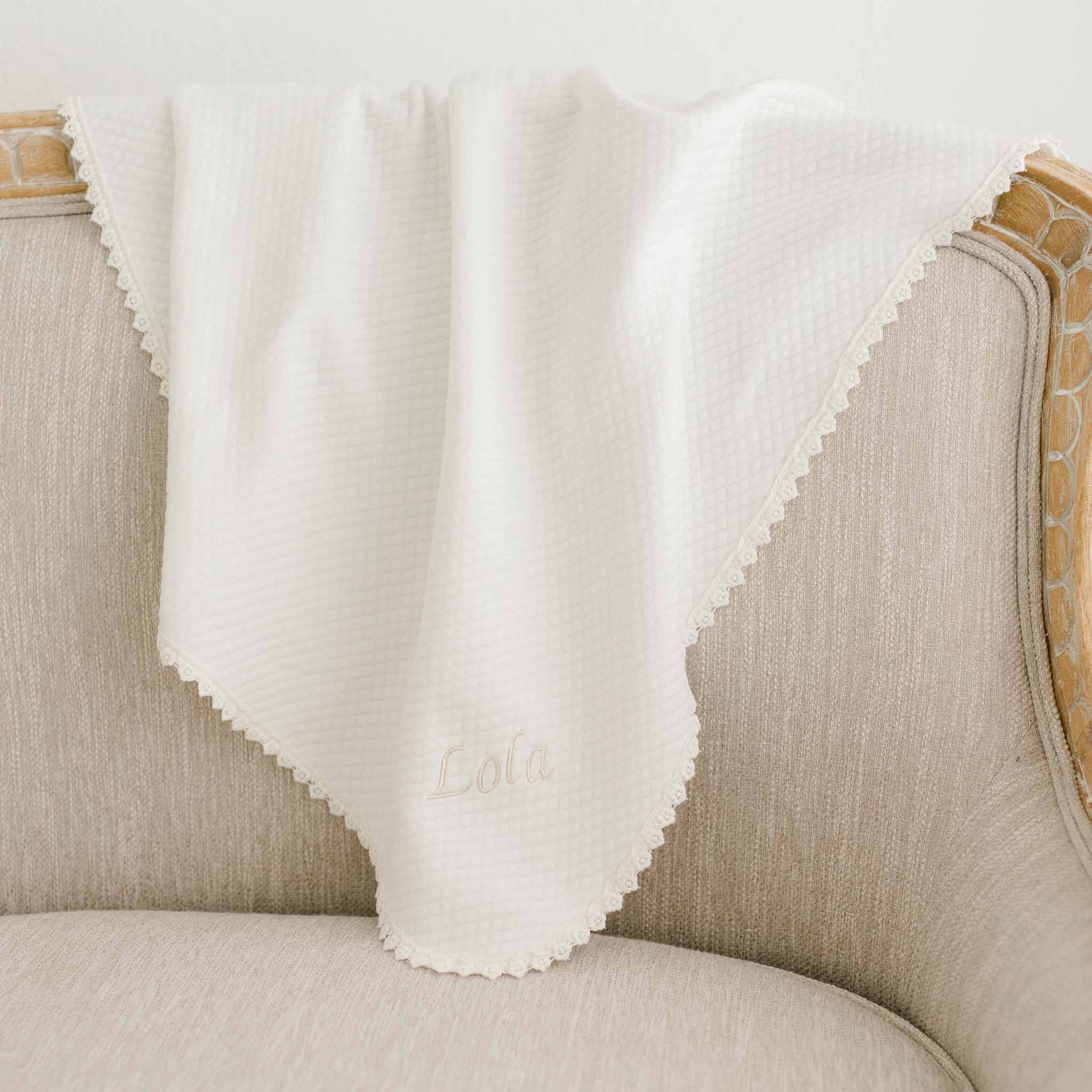 Lola Christening Personalized Blanket draped over a beige chair, made from soft light ivory cotton with delicate lace trim and 'Lola' embroidered on the corner, perfect for christening ceremonies.