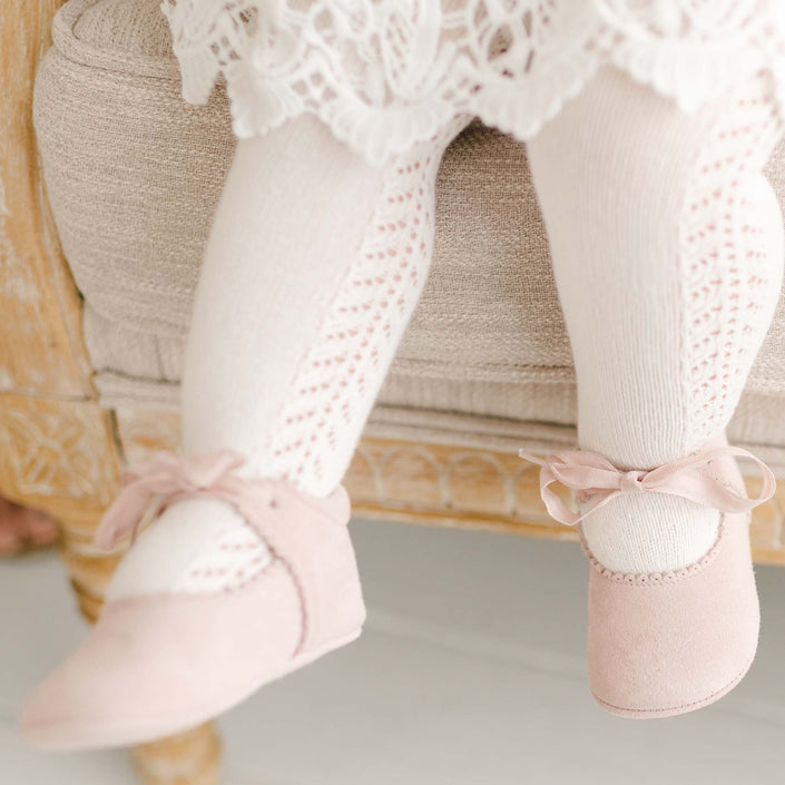 Close-up of a baby girl wearing the Lola Christening Suede Tie Mary Janes, crafted from soft rosy pink suede with scalloped edge detailing and tied with a delicate cotton ribbon, perfect for a christening outfit.