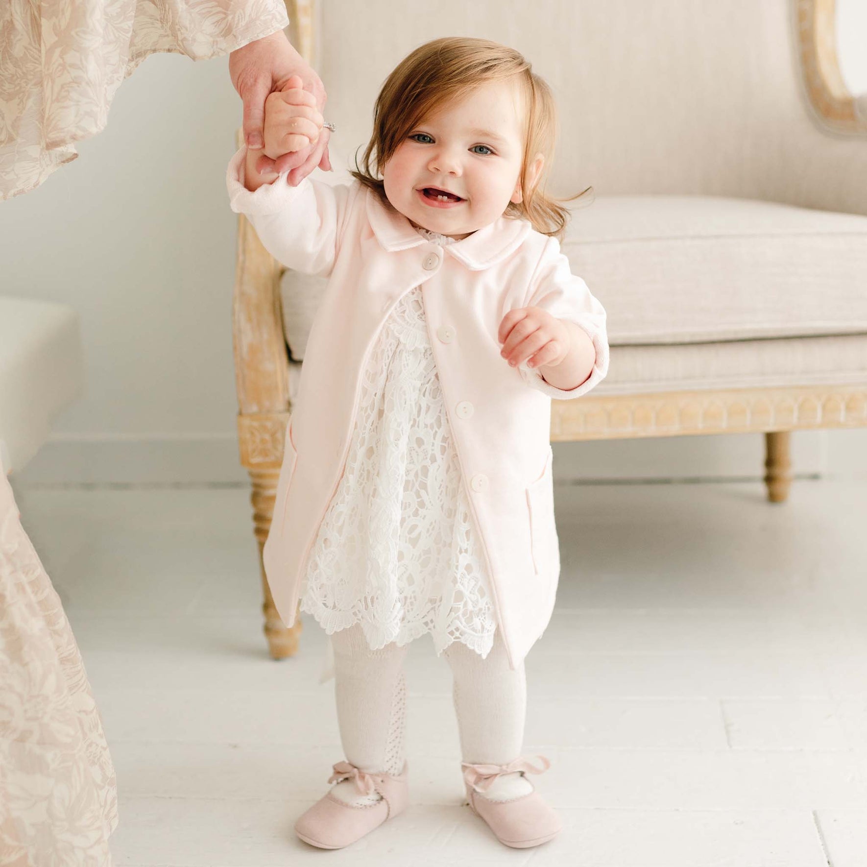 Baby girl wearing the Lola Christening Trench Coat in soft pink, layered over the Lola Christening Romper. The cotton French terry coat features a petite collar, button closures, and front pockets, while she holds an adult's hand and smiles.