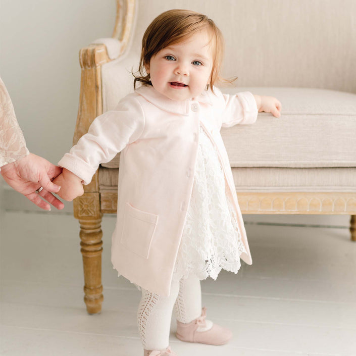 Baby girl wearing the Lola Christening Trench Coat over the Lola Christening Romper. The soft pink coat is made from cotton French terry and features a petite collar, button closures, and front pockets, perfect for layering over a christening outfit.