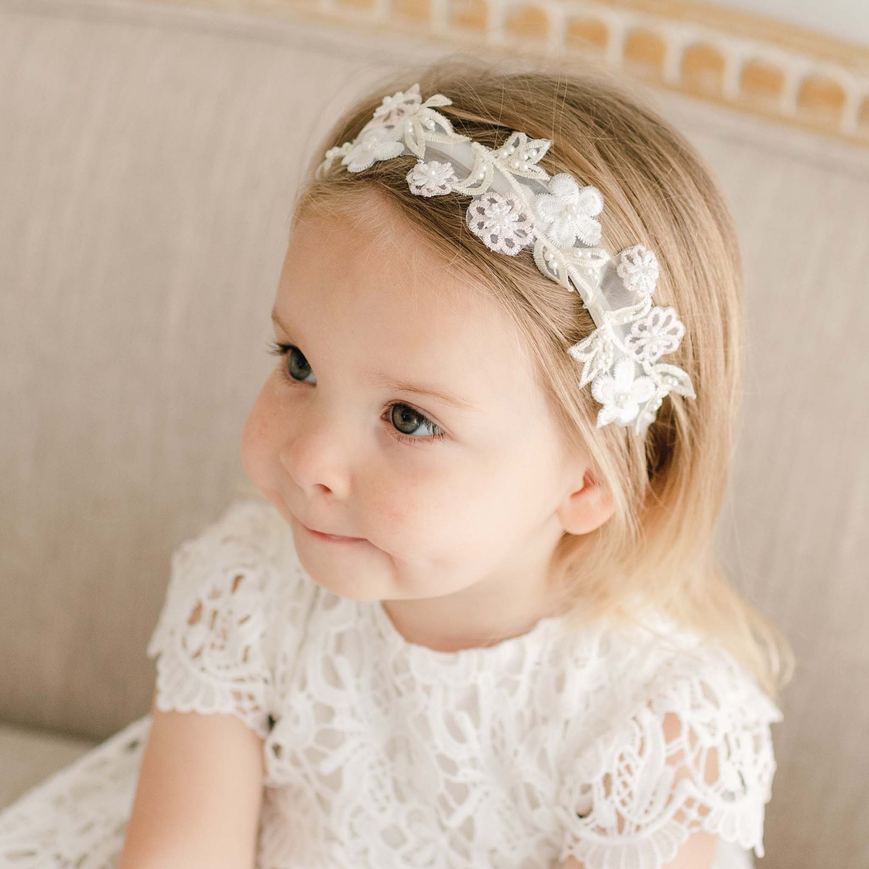 A toddler girl wearing the Lola Beaded Flower Headband, which features a velvet elastic band decorated with pastel pink and ivory flowers. She gazes sideways with her lips pursed.