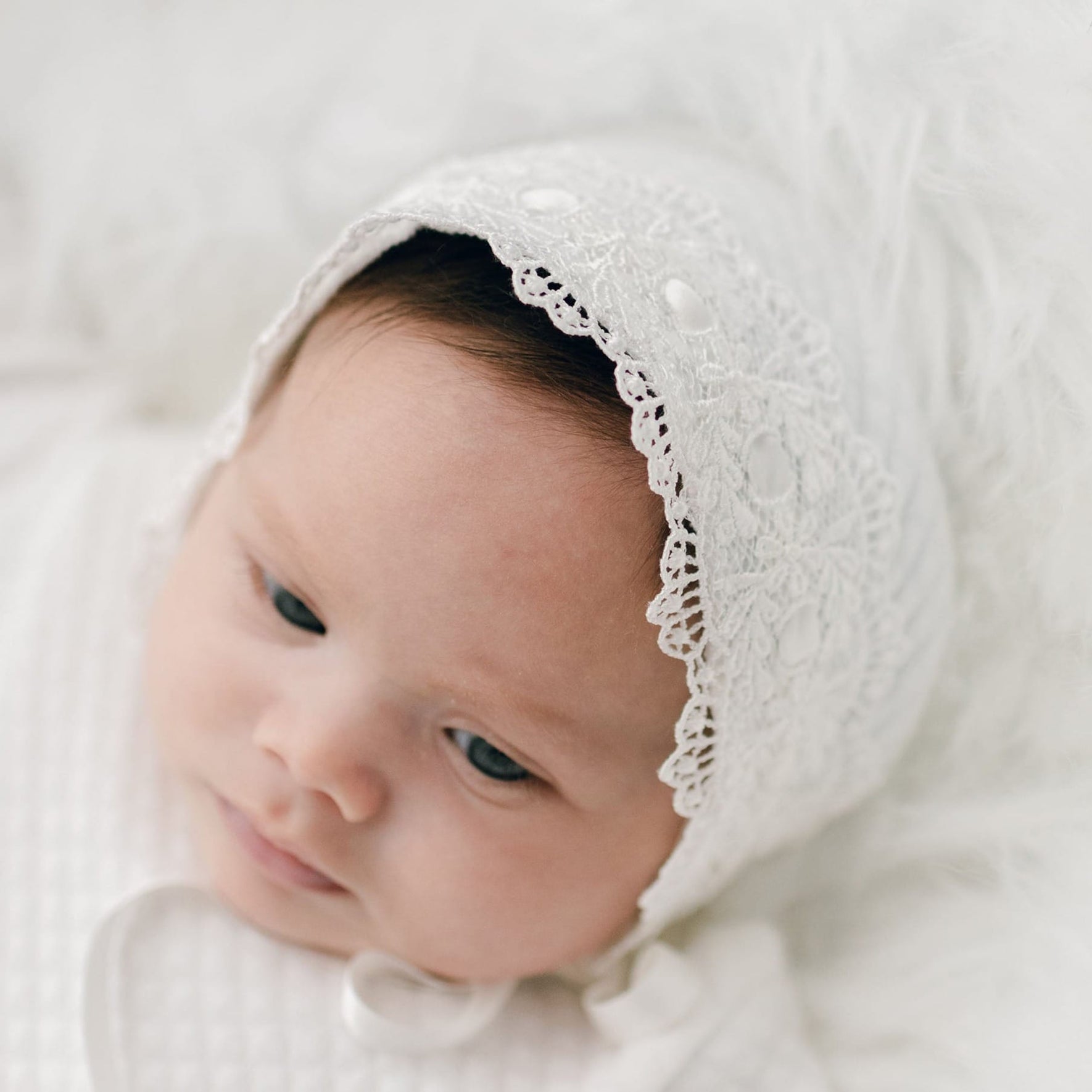 A newborn girl is lying down, looking off to the side. She is wearing a white lace Madeline Quilted Newborn Bonnet and rests against a soft, white background. The baby has a calm expression and is dressed in light-colored clothing made from quilted cotton.