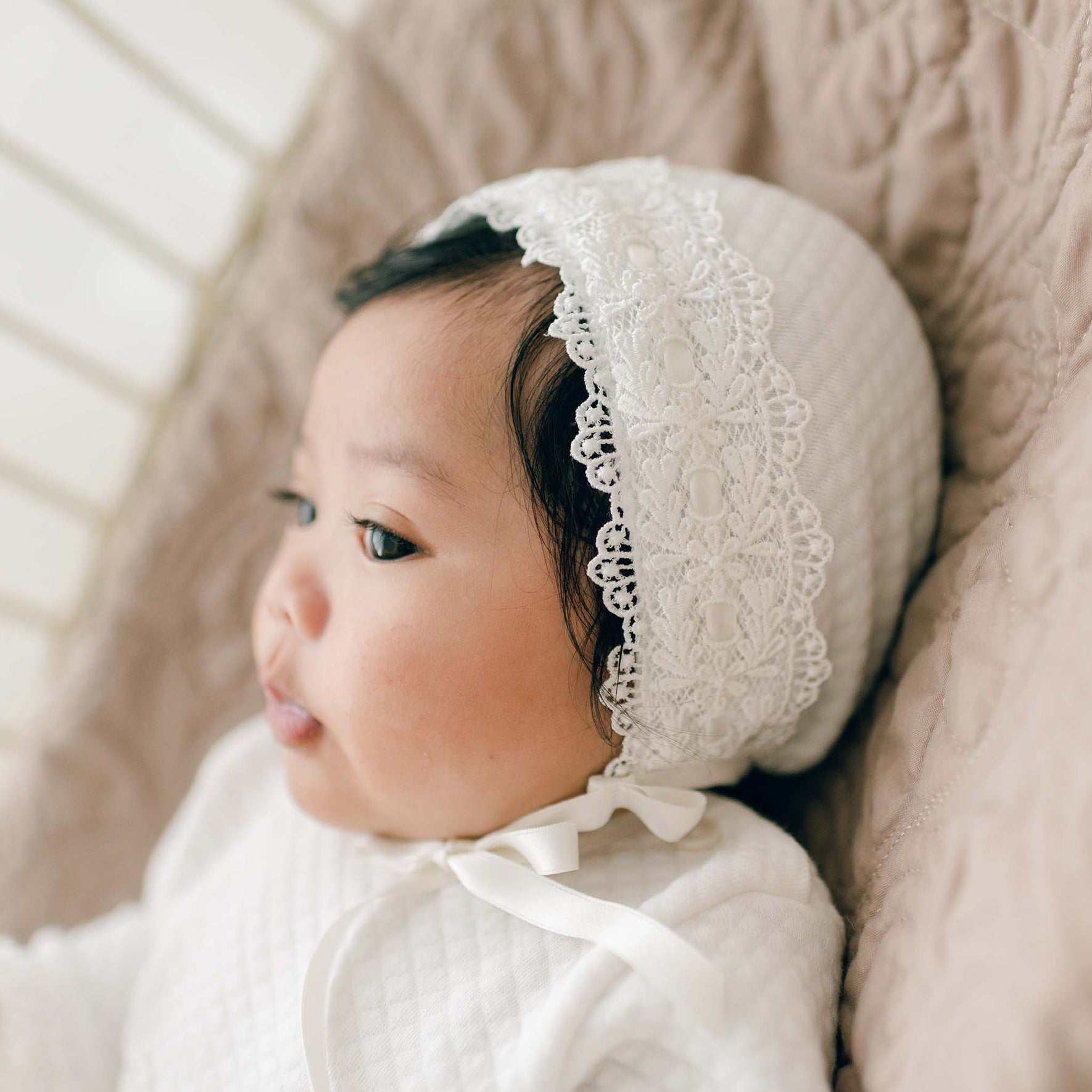 A newborn girl is lying down wearing a white outfit and a Madeline Quilted Newborn Bonnet. Viewed in profile, she rests on a cushioned surface with a quilted cotton background. The photo is captured in soft, natural lighting.