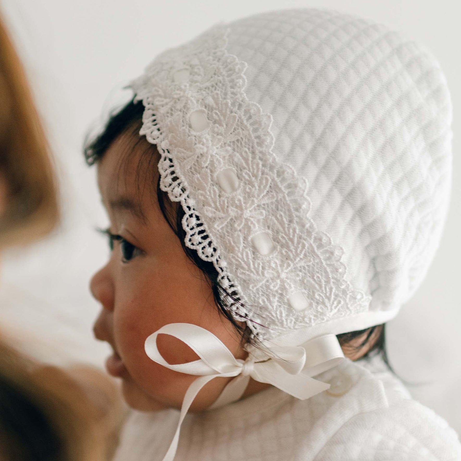 A newborn girl is wearing the Madeline Quilted Newborn Bonnet, a white textured piece with lace detailing along the edge and a satin ribbon tied under her chin. The bonnet, made from quilted cotton, is shown in intricate detail as the baby is seen from the side against a softly blurred, neutral background.