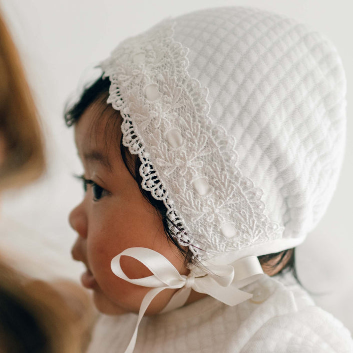 A newborn girl is wearing the Madeline Quilted Newborn Bonnet, a white textured piece with lace detailing along the edge and a satin ribbon tied under her chin. The bonnet, made from quilted cotton, is shown in intricate detail as the baby is seen from the side against a softly blurred, neutral background.