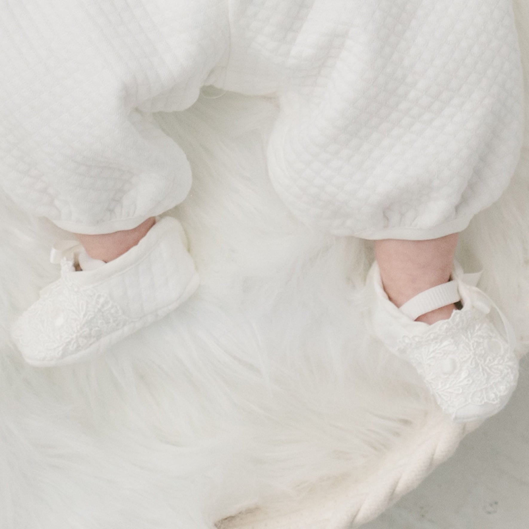 An infant wearing textured white pants and Madeline Quilted Booties is lying on a fuzzy white surface.