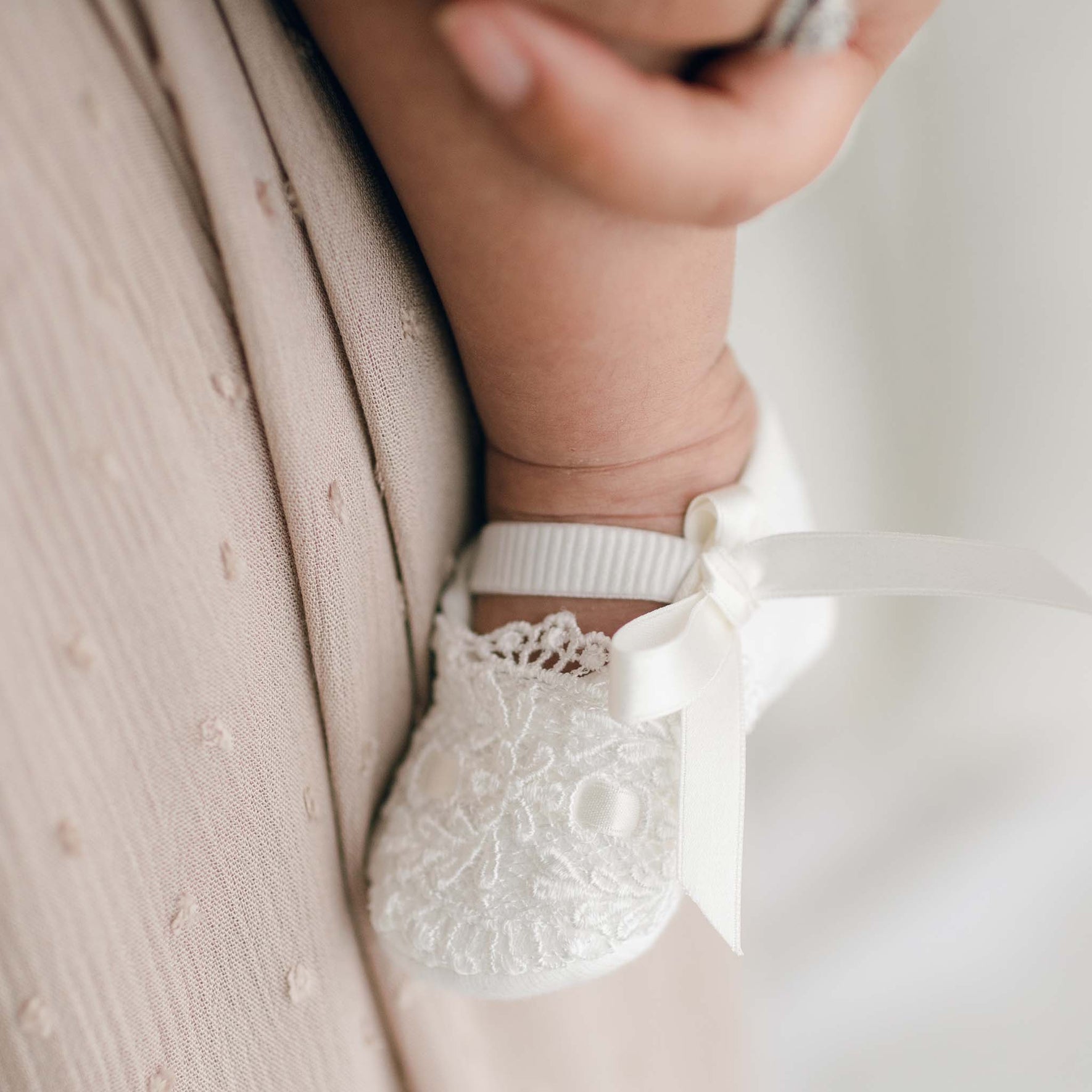 Close-up of a baby's foot wearing white, lace-trimmed Madeline Quilted Booties with small silk ribbon bows. The infant's foot is being gently held by an adult's hand, visible in the background, resting on a pale, textured fabric.