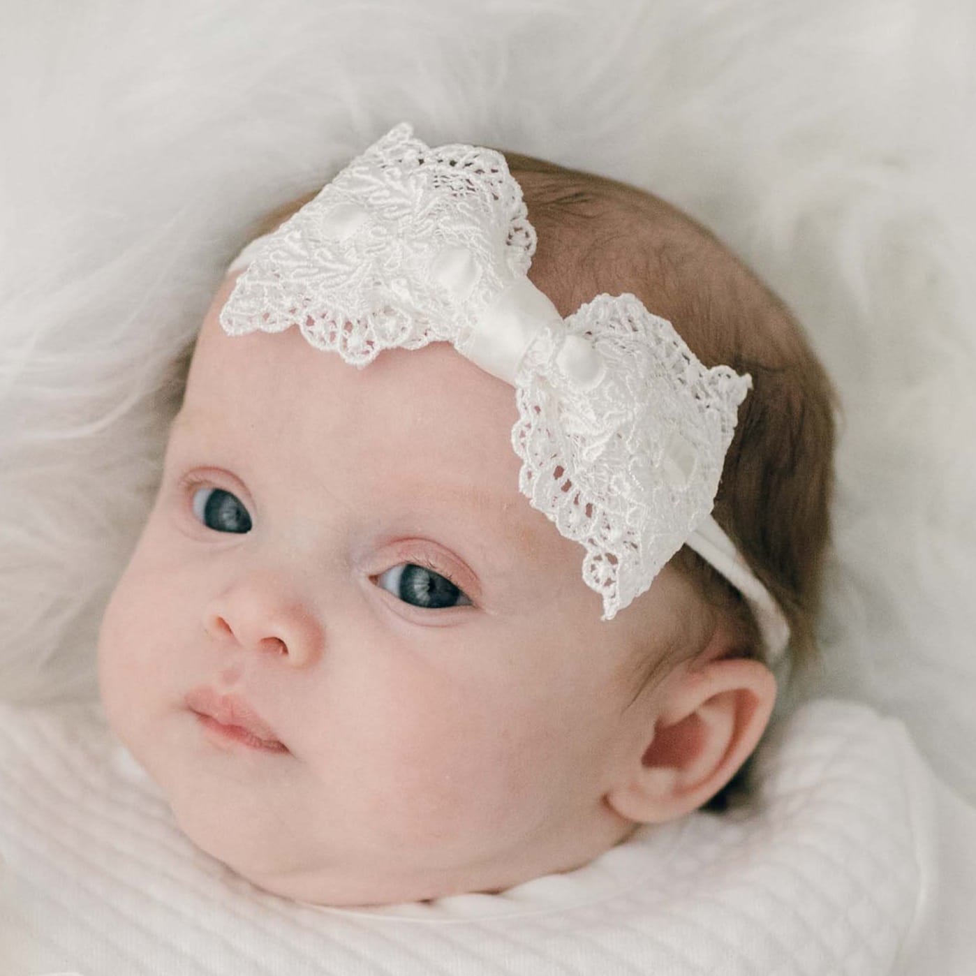 A baby with light skin and blue eyes is lying down on a white surface. The baby is wearing a white outfit and an ivory Madeline Lace Bow Headband. The baby looks directly at the camera with a calm expression.