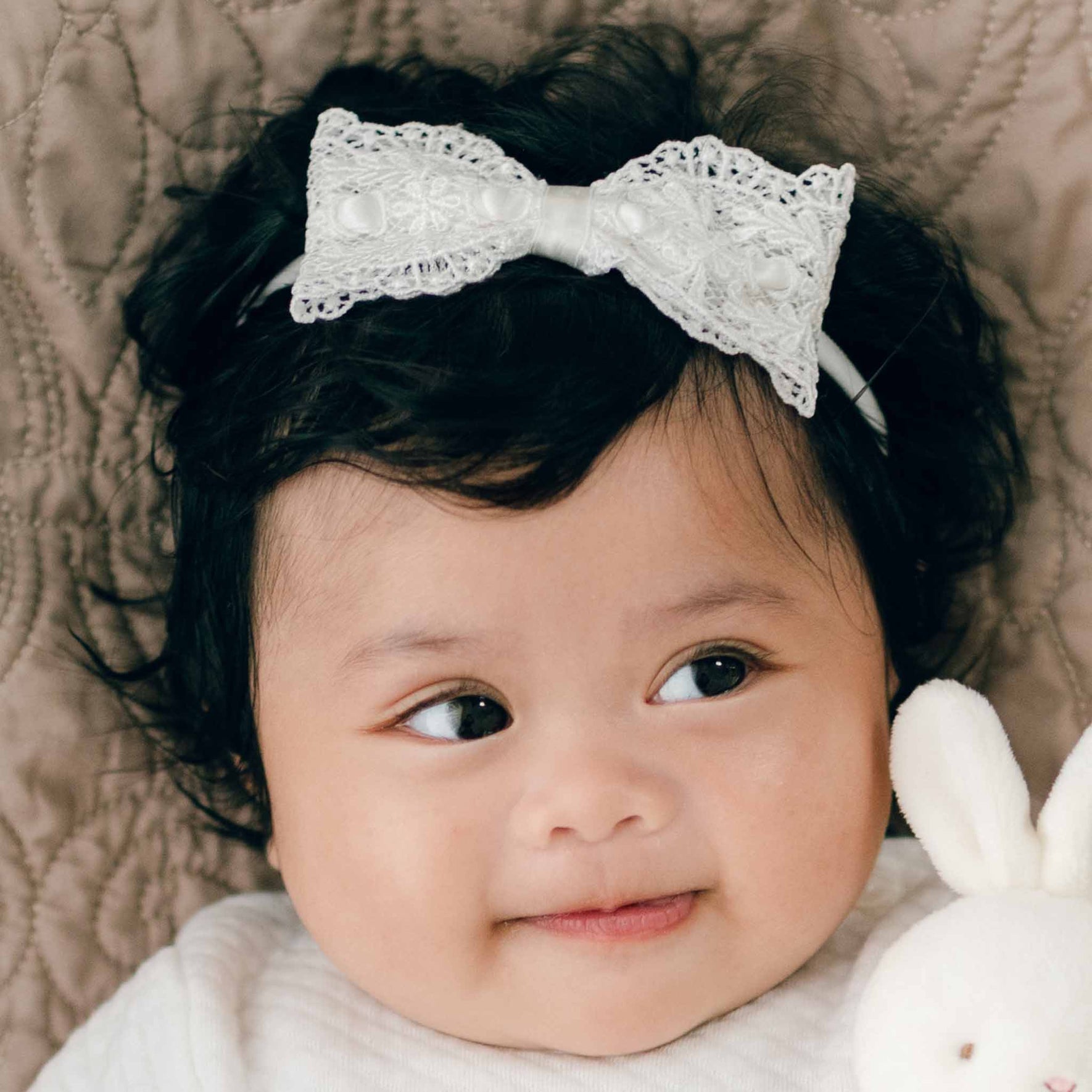 A baby with dark hair and a Madeline Lace Bow Headband is lying down on a soft, textured surface, looking to the side with a slight smile. The baby is also holding a white plush toy.