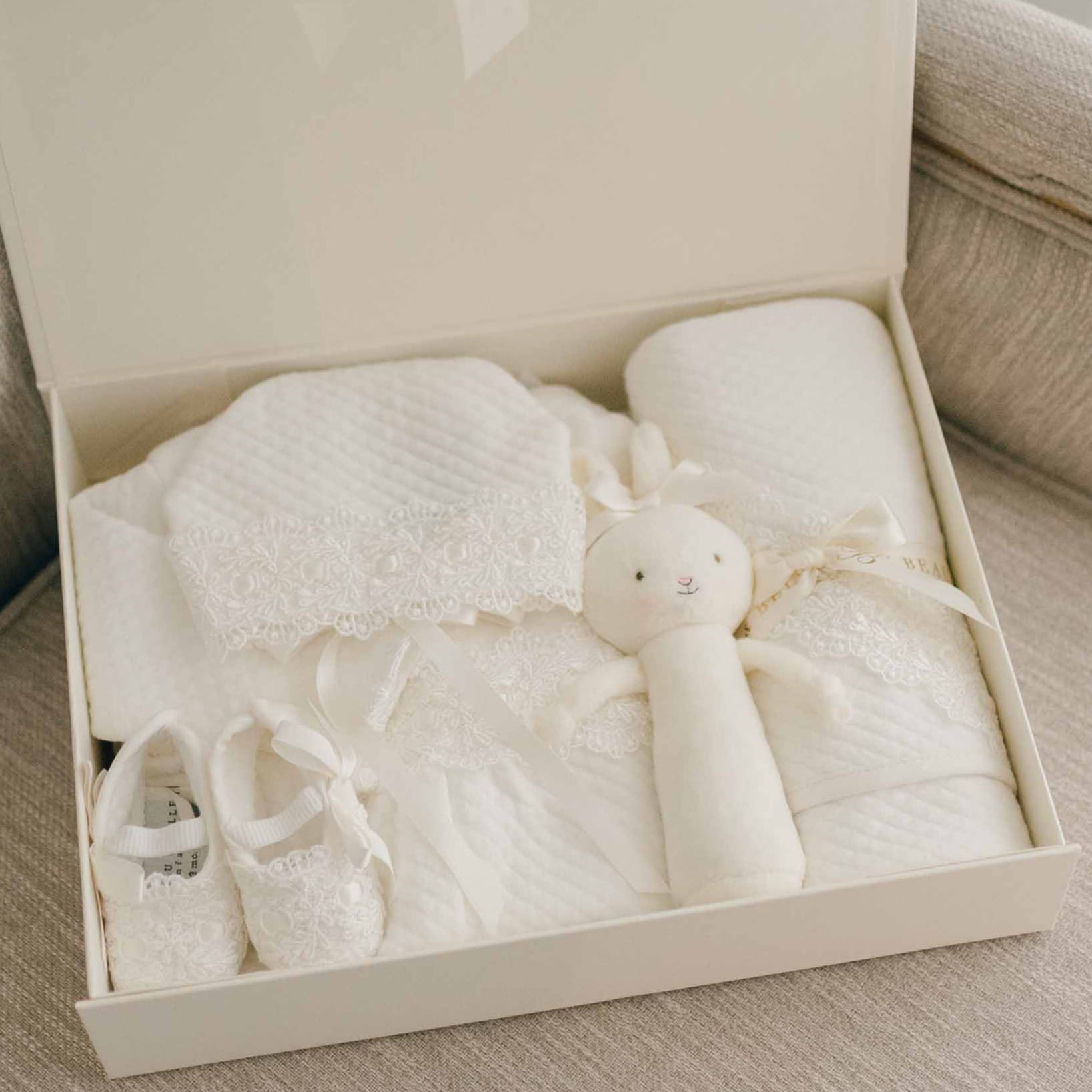 The Madeline Newborn Gift Set, now with a 10% discount, elegantly displays a take-home outfit presentation that includes baby clothes, a chic Madeline romper, lace-detailed shoes, and an adorable bunny-shaped soft toy. This designer ensemble is finished with a textured blanket in a creamy white palette set against a light-colored backdrop.