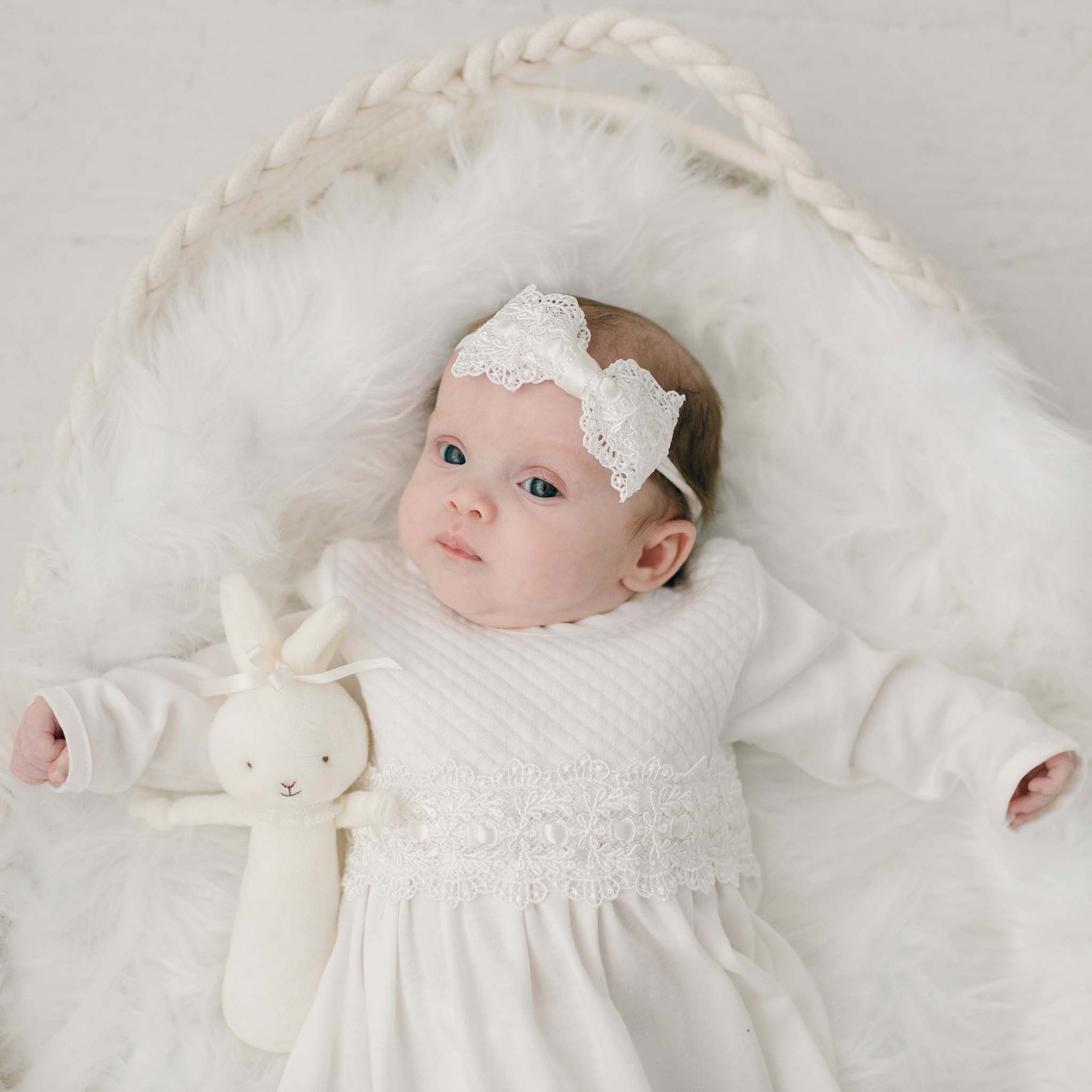 A baby is lying in a white, fuzzy blanket. The baby is wearing a white headband with lace and a Madeline Newborn Gown adorned with ivory lace detail. The baby holds a small, white stuffed bunny. The background is light and neutral.