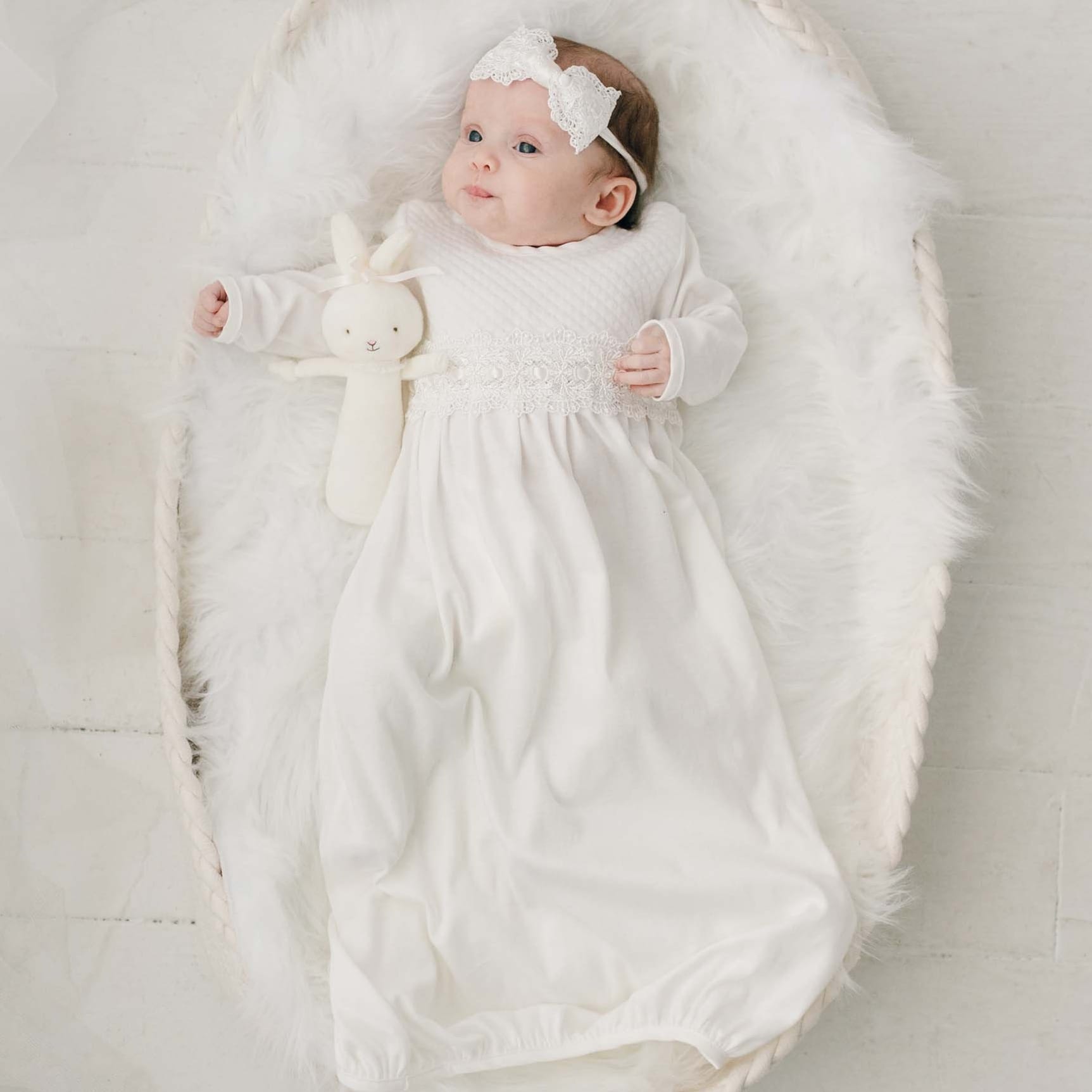 A baby is lying on a fluffy white surface in a wicker basket. She is dressed in a long, ivory lace Madeline Newborn Gown made of soft pima cotton with delicate detailing and a matching headband. Holding a white stuffed bunny, the baby's gaze is directed upwards.