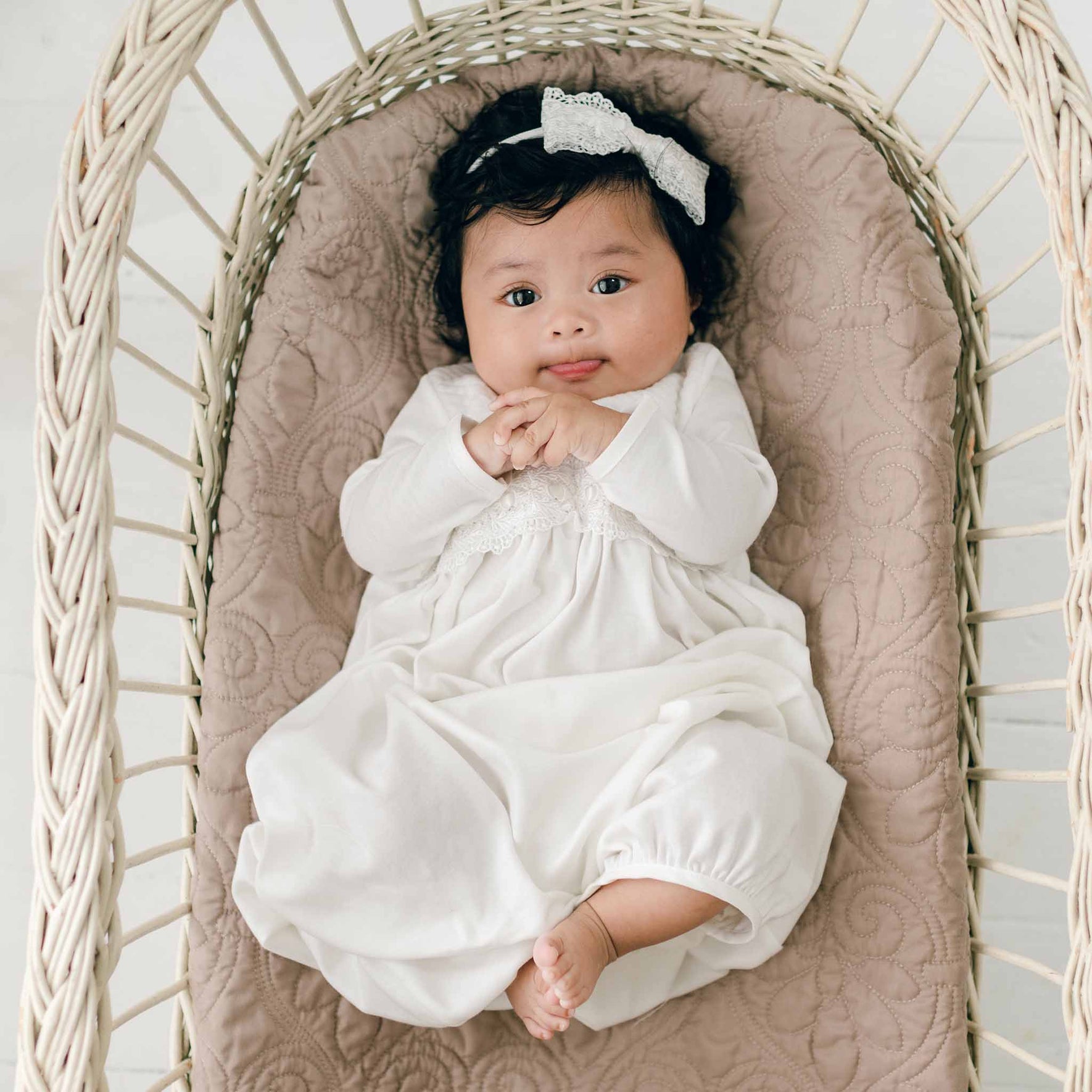 A baby wearing a Madeline Newborn Gown and an ivory lace bow headband lies on a cushioned surface in a wicker basket. The baby has dark hair, wide eyes, and is looking up while slightly smiling with clasped hands.