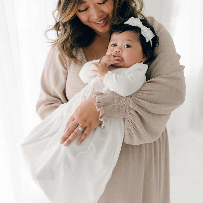 A woman with medium-length wavy hair is holding a baby dressed in a Madeline Newborn Gown adorned with ivory lace. The baby, in their pima cotton outfit and white headband, is sucking on their thumb while the woman smiles and looks down at the child. Both are dressed in light-colored clothing.
