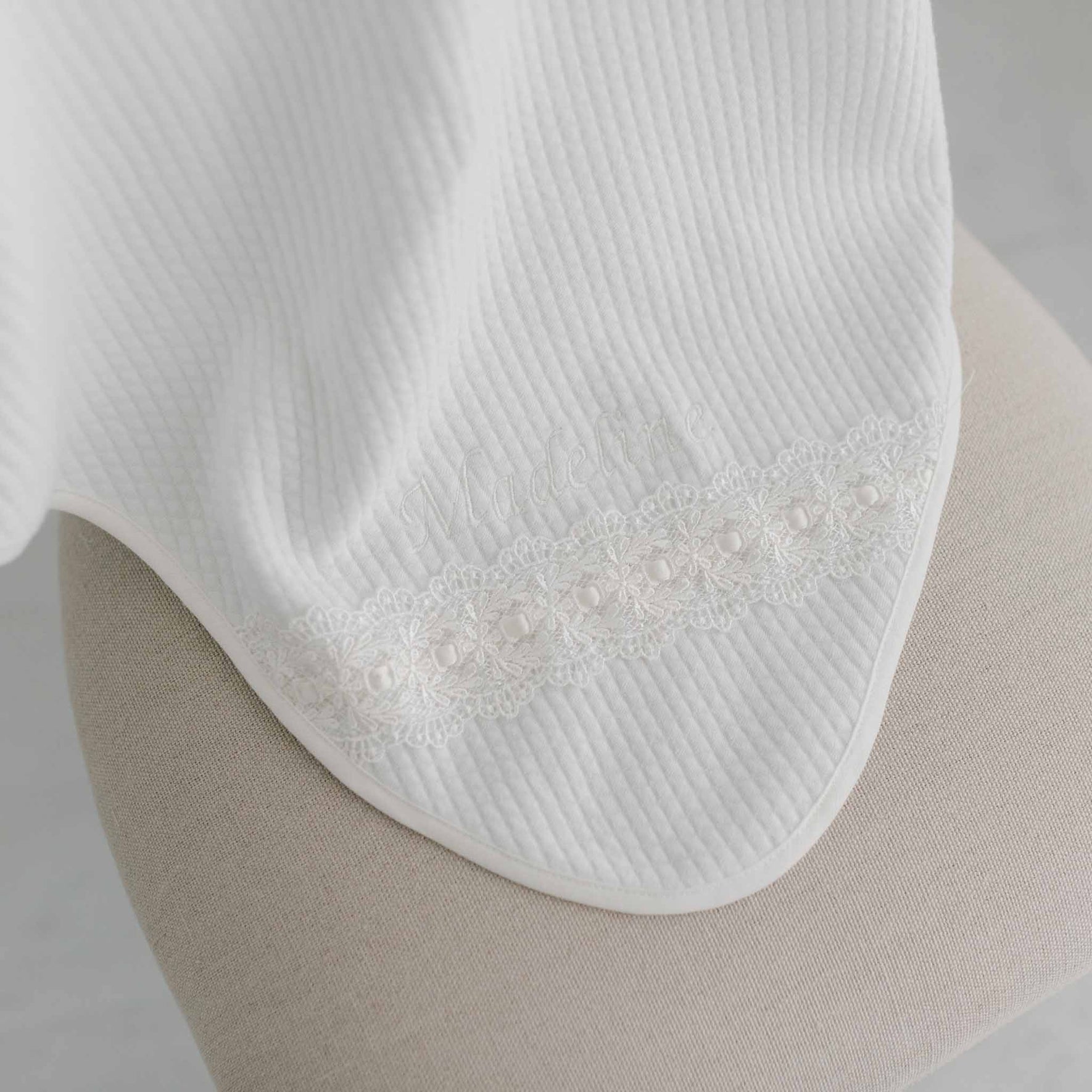 A close-up of a white textured fabric draped over a beige surface features the Madeline Personalized Blanket, highlighting its delicate ivory lace with floral patterns and subtle custom embroidery spelling "Madeleine.
