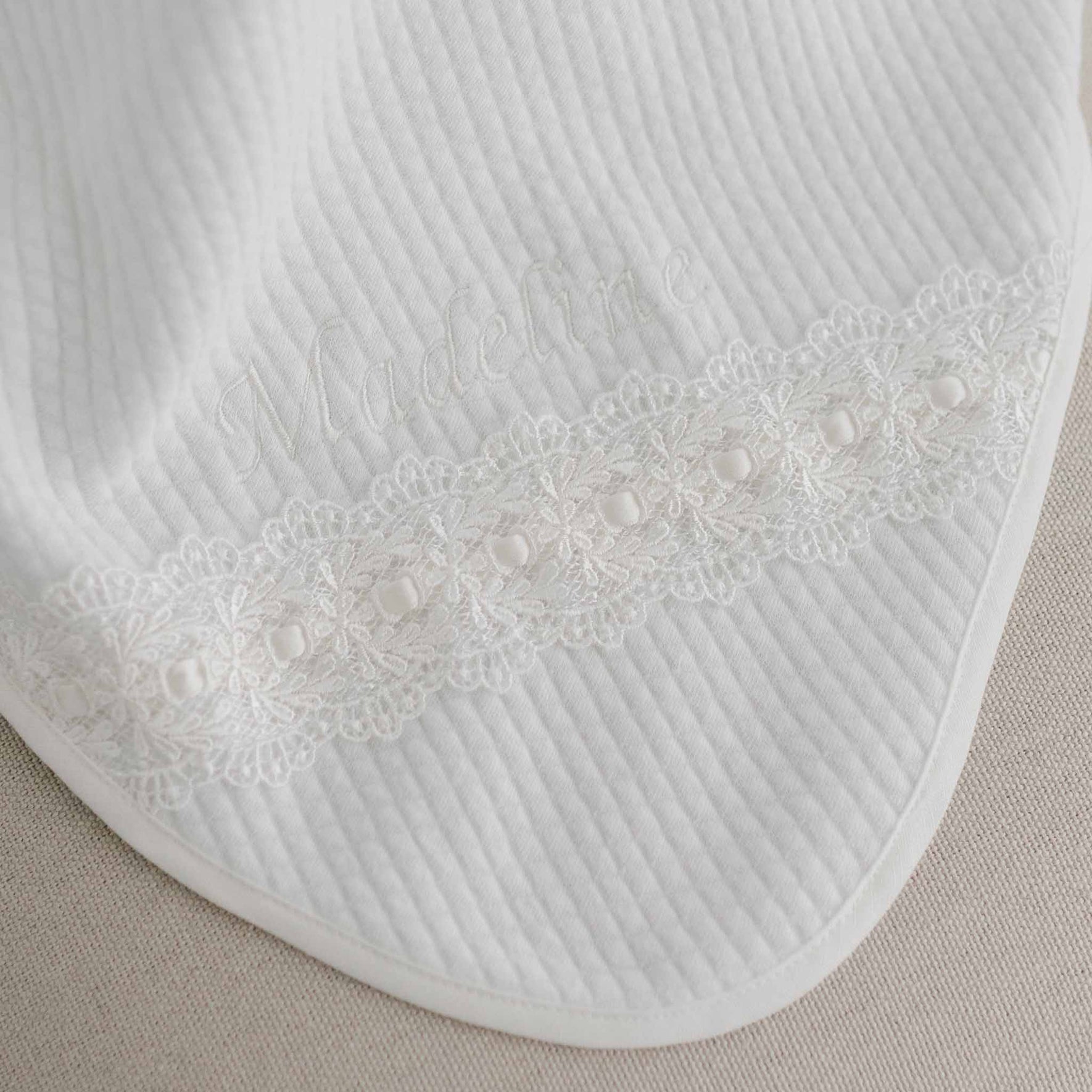 A detailed view of the Madeline Personalized Blanket, featuring intricate ivory lace detailing and the name "Madeline" expertly embroidered near the edge. The texture of the fabric is visible, highlighting its custom embroidery as it lays flat on a neutral background.