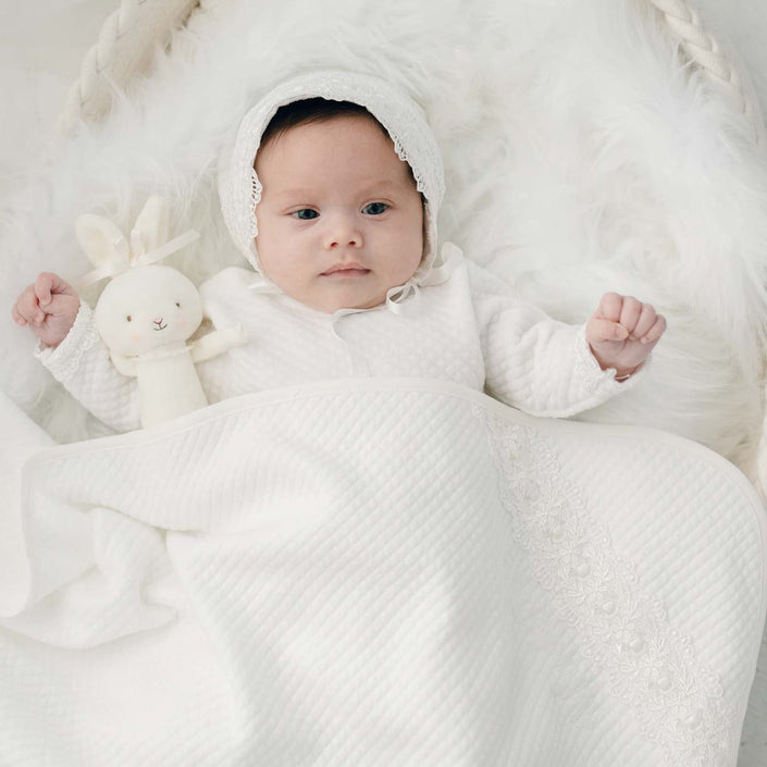 A baby wearing a white outfit and bonnet lies on a soft, fluffy surface, holding a cream-colored stuffed animal. The Madeline Personalized Blanket, an exquisite quilted cotton blanket with delicate custom embroidery, partially covers the baby.