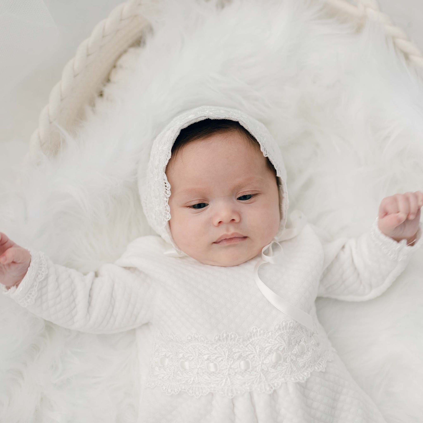 A baby wearing a white outfit and a lace bonnet lies on a fluffy white surface. The Madeline Quilted Newborn Romper, handmade in the USA, adds an extra touch of charm. The baby has a calm expression and is surrounded by soft, textured materials.