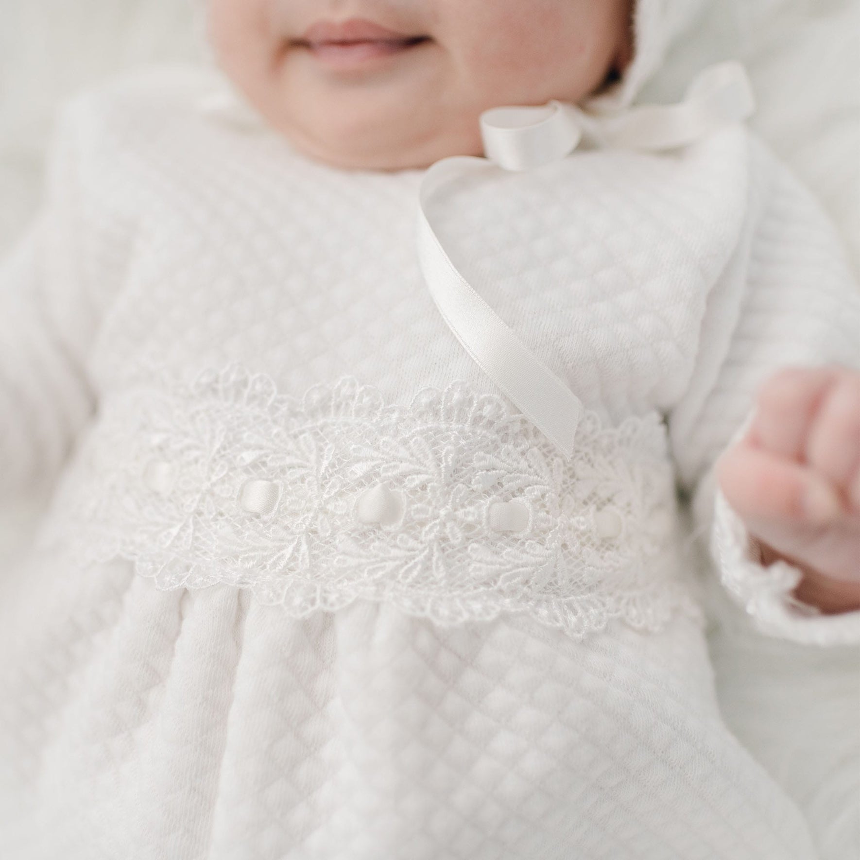 A close-up image of an infant wearing a white, quilted cotton outfit adorned with lace and ribbon details. The baby’s face is partially visible, and one hand is raised and slightly blurred in the foreground. The focus is on the intricate lace and ribbon on the Madeline Quilted Newborn Romper.