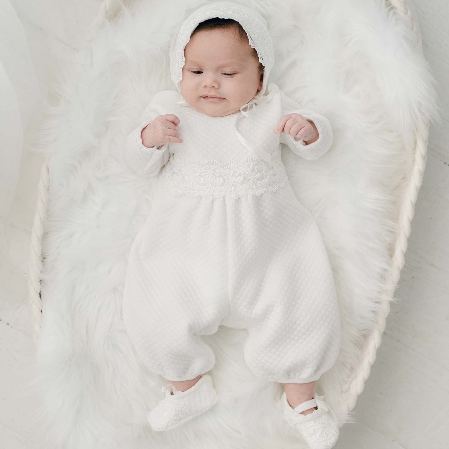 A baby lies on a white, fluffy blanket in a basket. The baby is wearing a Madeline Quilted Newborn Romper with a lace bonnet and matching booties, all handmade in USA. The baby's hands are slightly raised as they look upward.