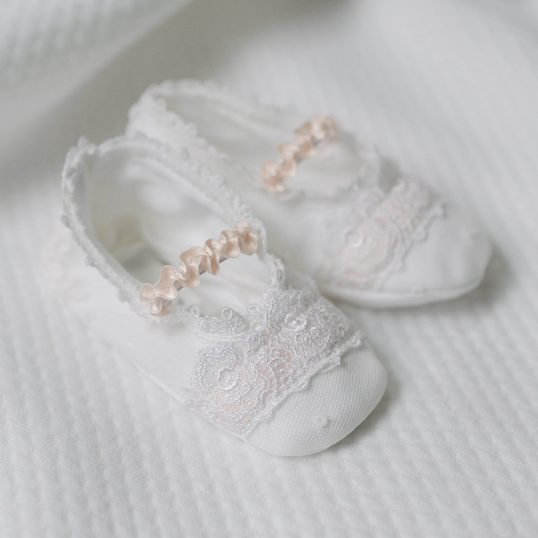 Flat lay photo of the Melissa Booties with lace details and pink ribbons on a white quilt surface. Close up details feature dotted Swiss and floral embroidered pattern netting overlay and white Venice lace covering the top. An accessory to luxury baby clothes. 