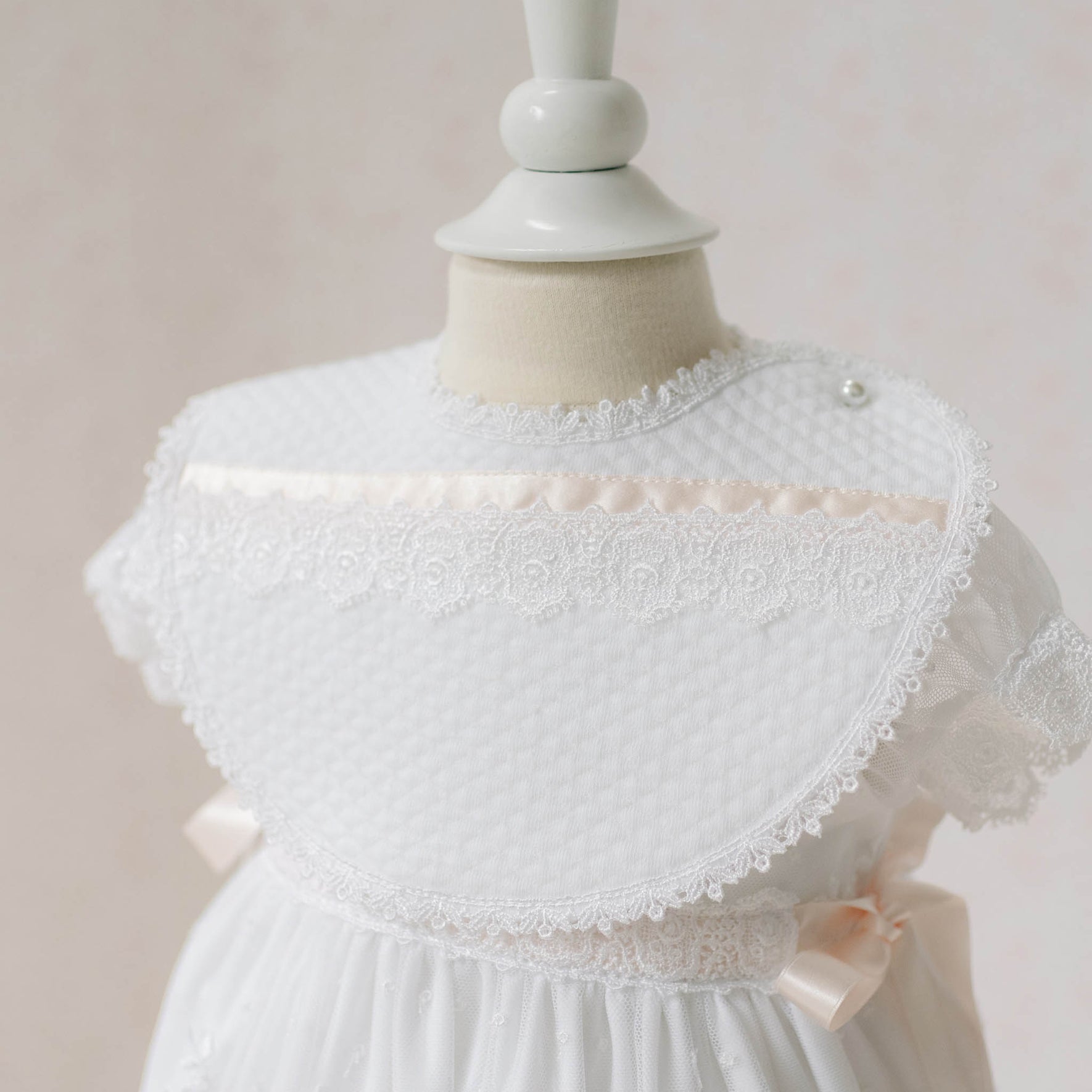 Close up photo of the Melissa Bib on a mannequin against a plain background. Details show white quilted textured cotton with Venice lace and silk ribbon. Perfect baptism gift from godparents.