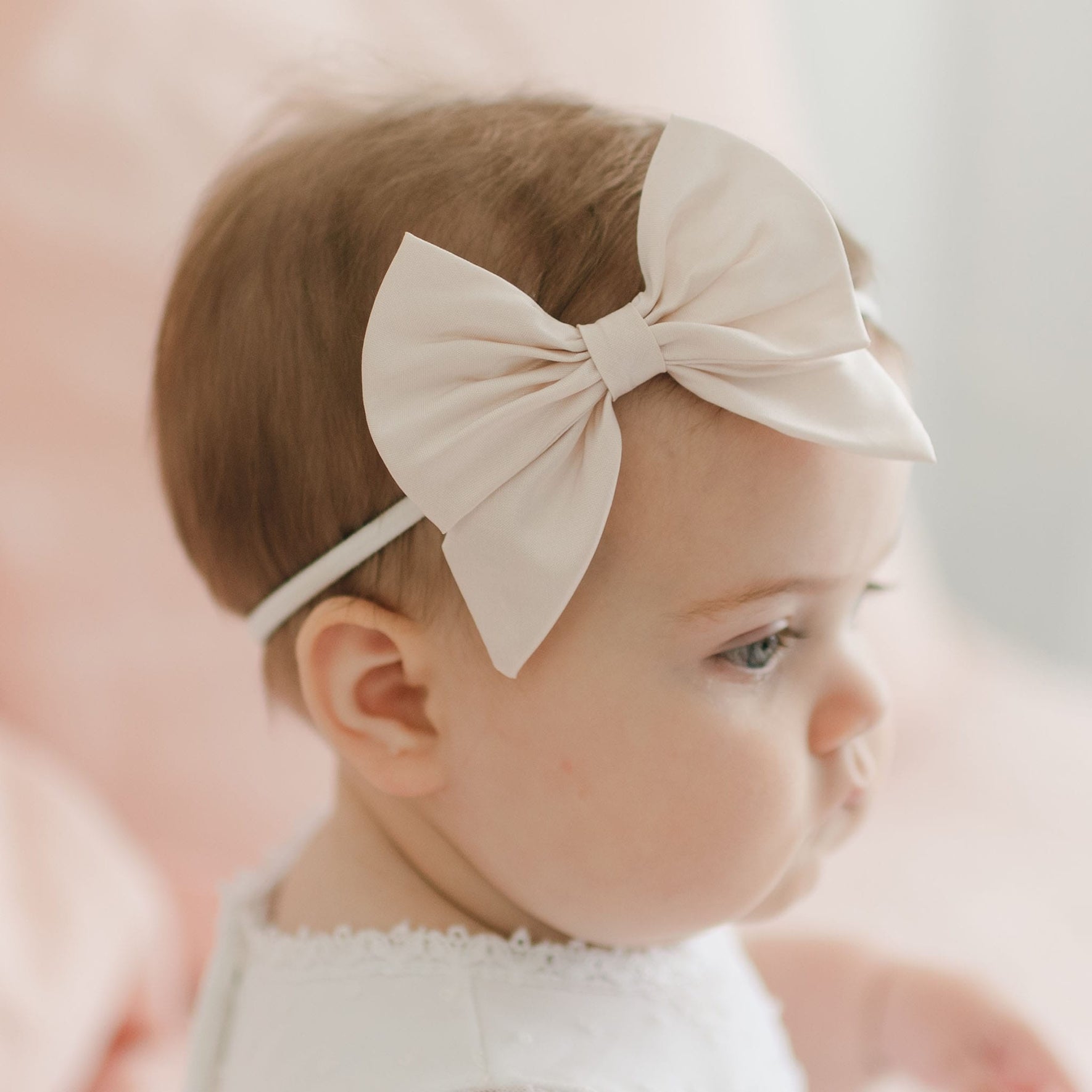 A baby wearing a white headband with a large bow is looking to the side. The baby has short brown hair and is dressed in a white outfit. The background is softly blurred with a light pink tone, complementing the Melissa Bow Headband featuring stretchy nylon for comfort.