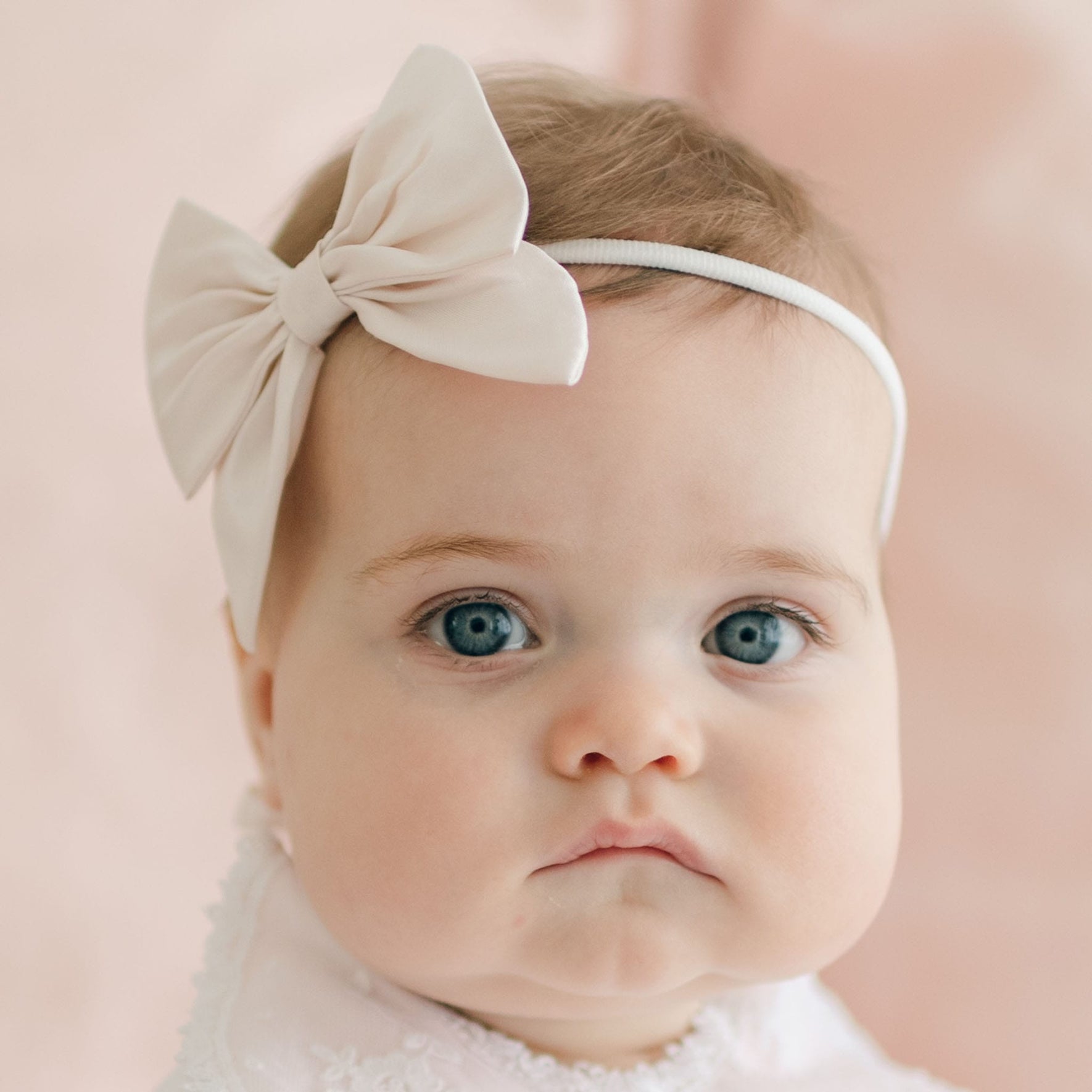 A baby with light-colored hair and blue eyes is wearing a white **Melissa Bow Headband** with a large pink silk ribbon bow. The baby is dressed in a light-colored outfit and has a neutral expression on their face. The background is a soft, light pink.