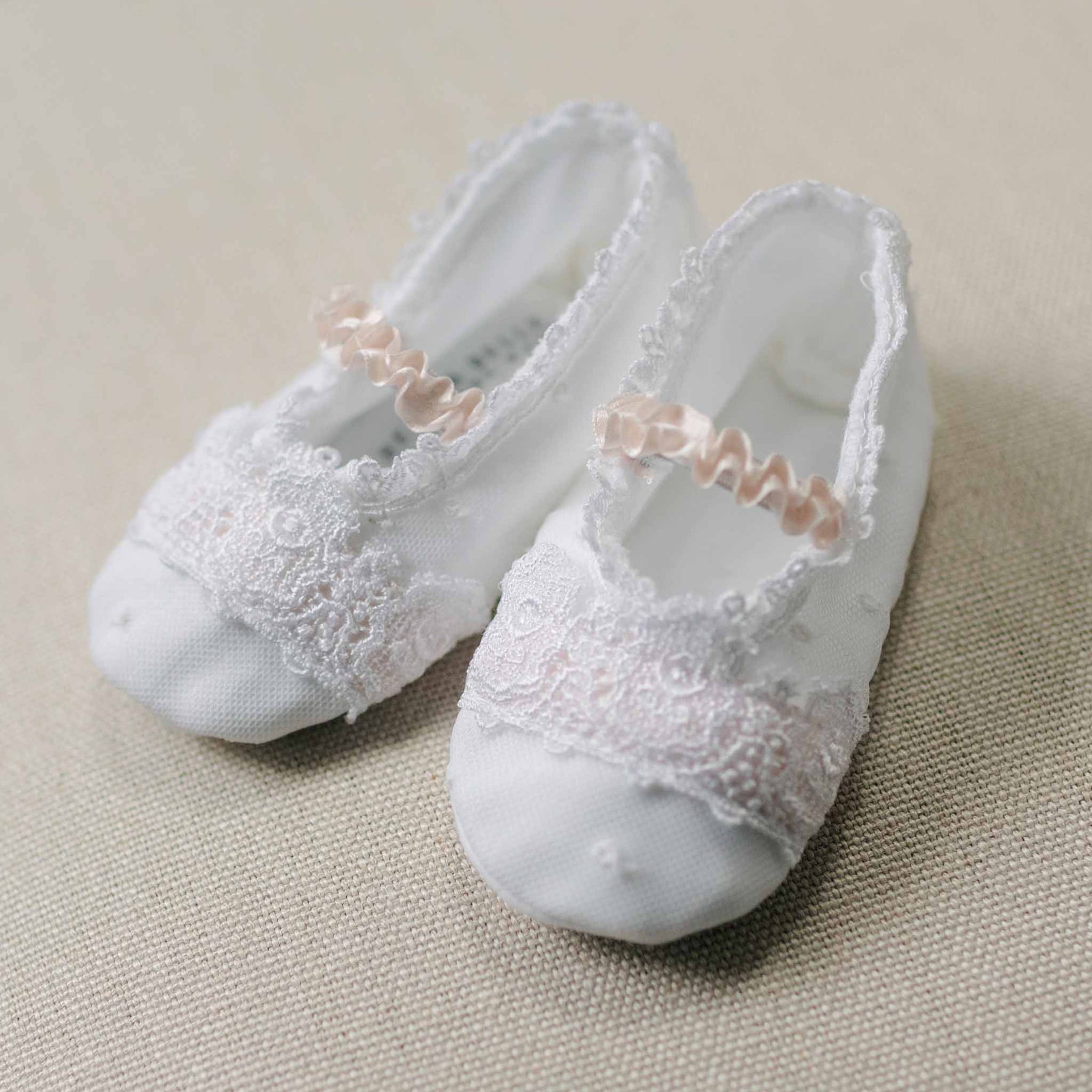 Flat lay photo of the Melissa Booties with lace details and pink ribbons on a beige linen surface. Close up details feature dotted Swiss and floral embroidered pattern netting overlay and white Venice lace covering the top. An accessory to high end baby clothes. 