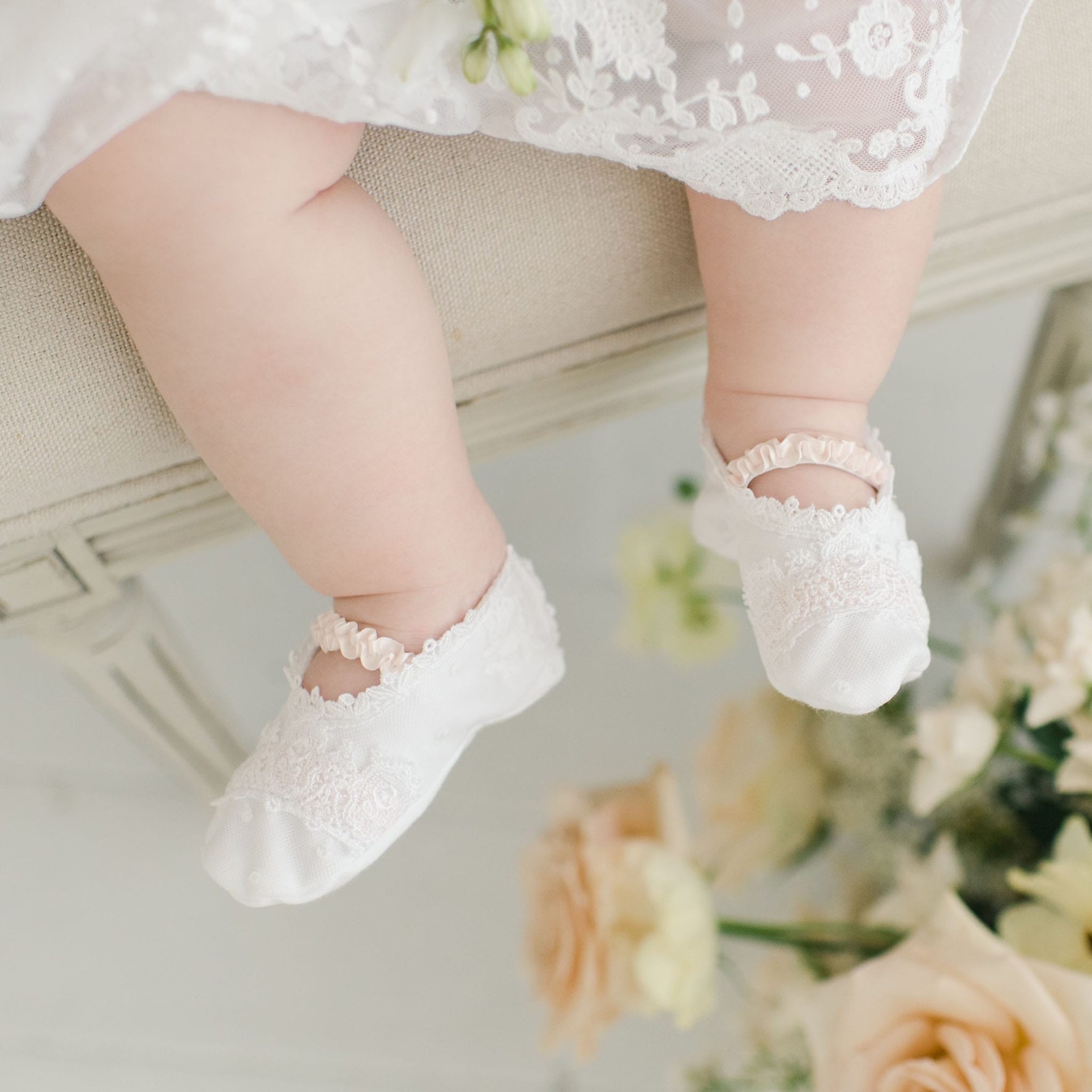 Close-up of a baby's legs wearing Melissa Christening Booties crafted from white Venice lace, seated on a light-colored surface. The baby is dressed in an elegant outfit made from 100% pima cotton. In the background, there are light-colored flowers, mainly white and soft peach roses.