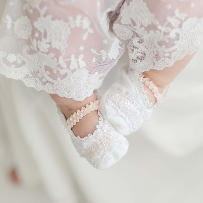 Close-up of a baby's feet wearing Melissa Christening Booties crafted from white Venice lace and pants with lace detailing. The background is a soft blur, putting focus on the intricate patterns and the baby's tiny feet.